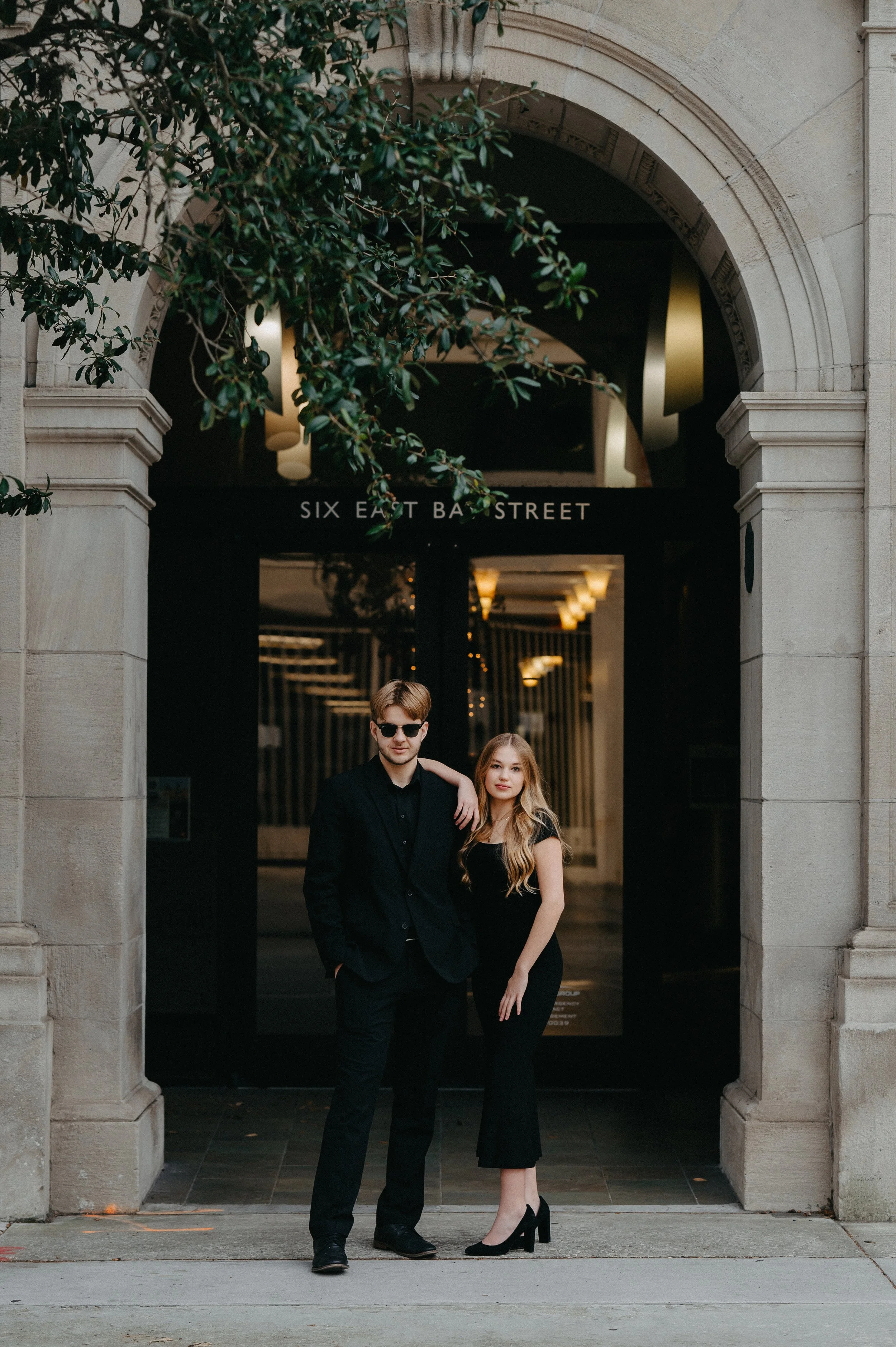 A young man and woman dressed in black standing in front of a building with an arched entrance labeled 'Six East Ba Street'. The man has sunglasses and a black suit, while the woman has long blonde hair and a black dress. She has her arm around his shoulder.