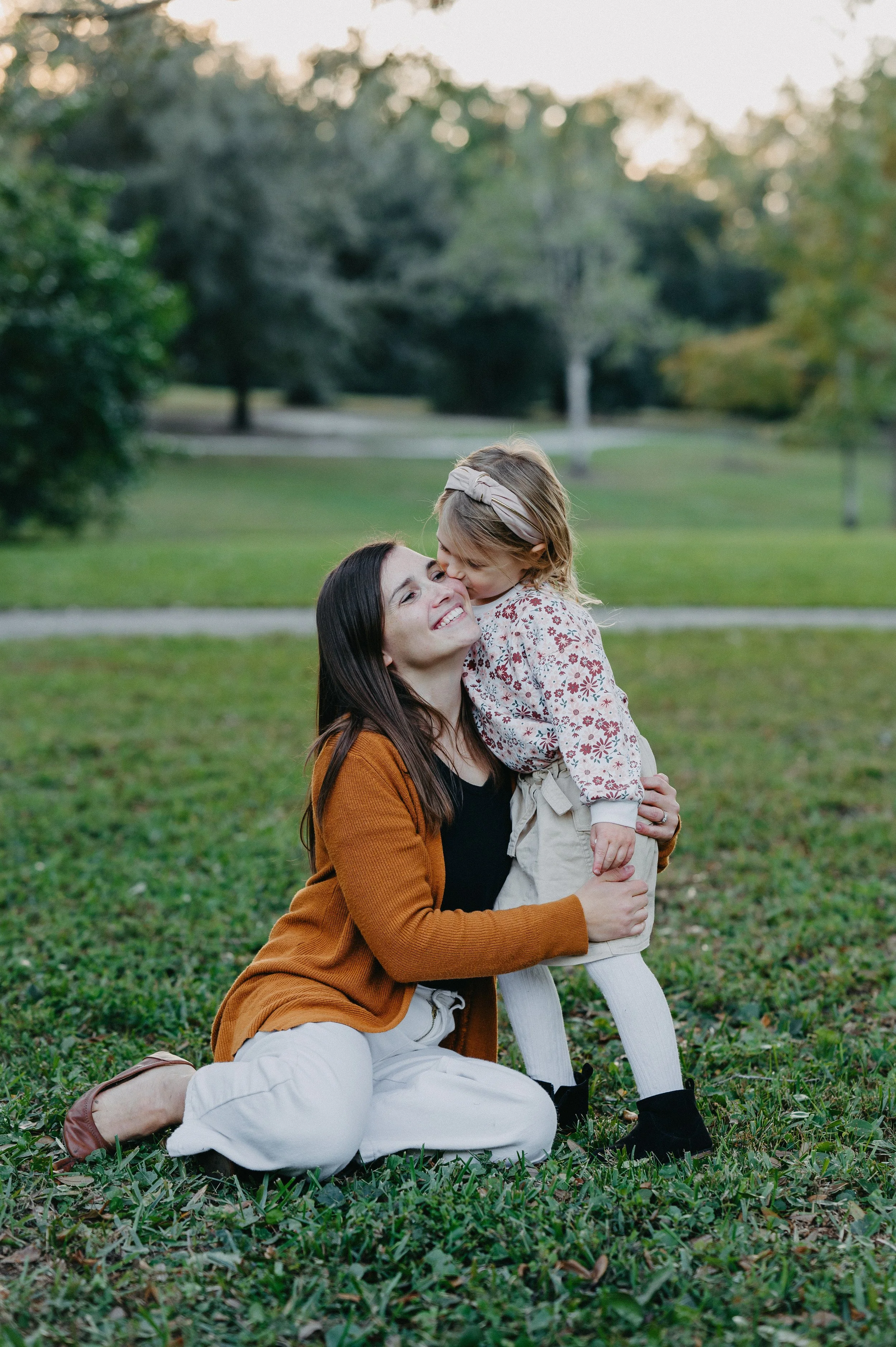 A woman and a young girl share a joyful moment outdoors in a park, with the woman kneeling on the grass and the girl standing in front of her. The girl is giving the woman a kiss on the cheek, and both are smiling.