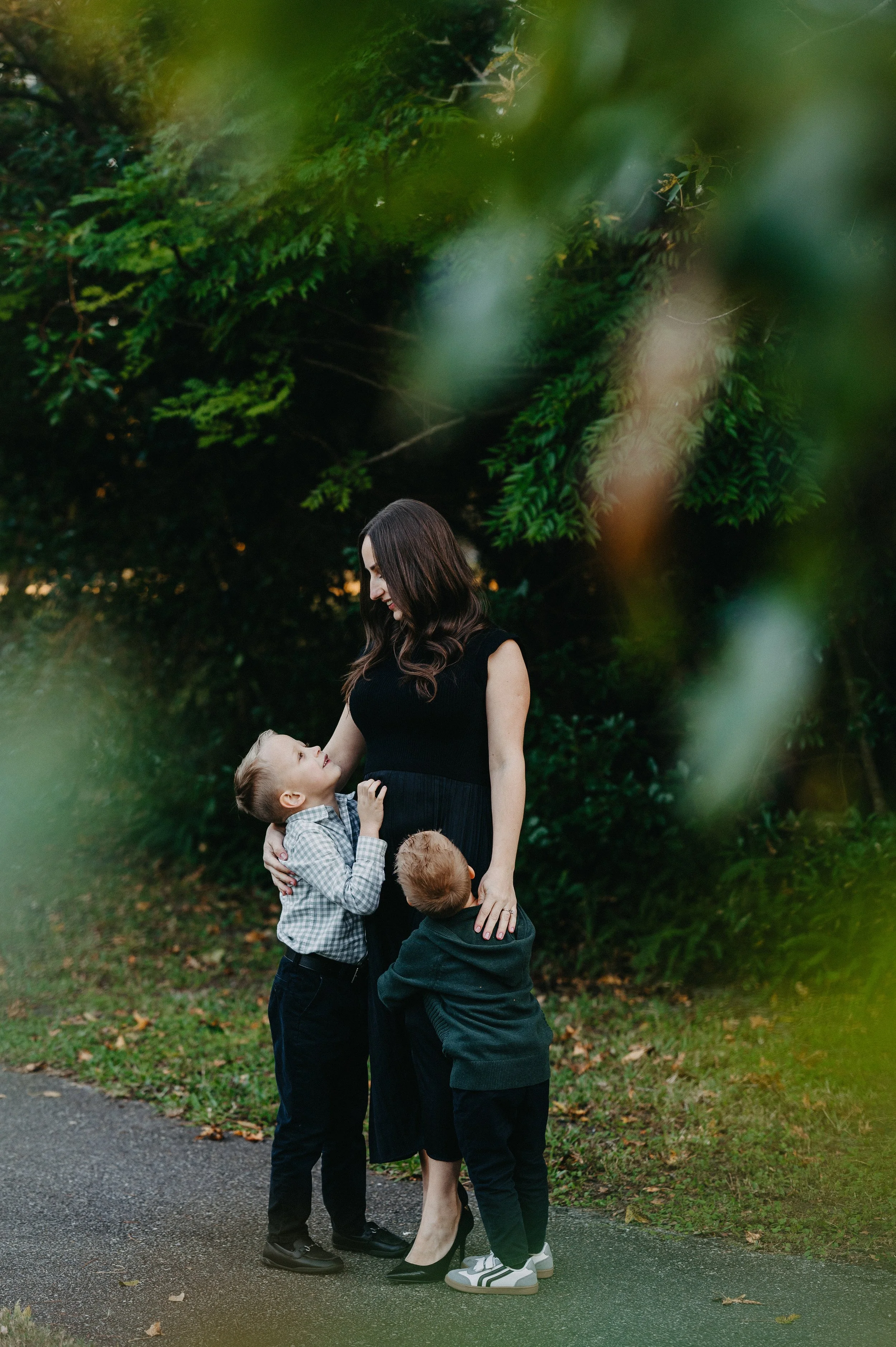 A woman with long dark hair, dressed in a black dress and heels, standing outdoors on a paved path, is being affectionately hugged by two young boys. The boys have light hair, one blond and one redhead, and are wearing casual clothing. They are in a lush, green, wooded area.