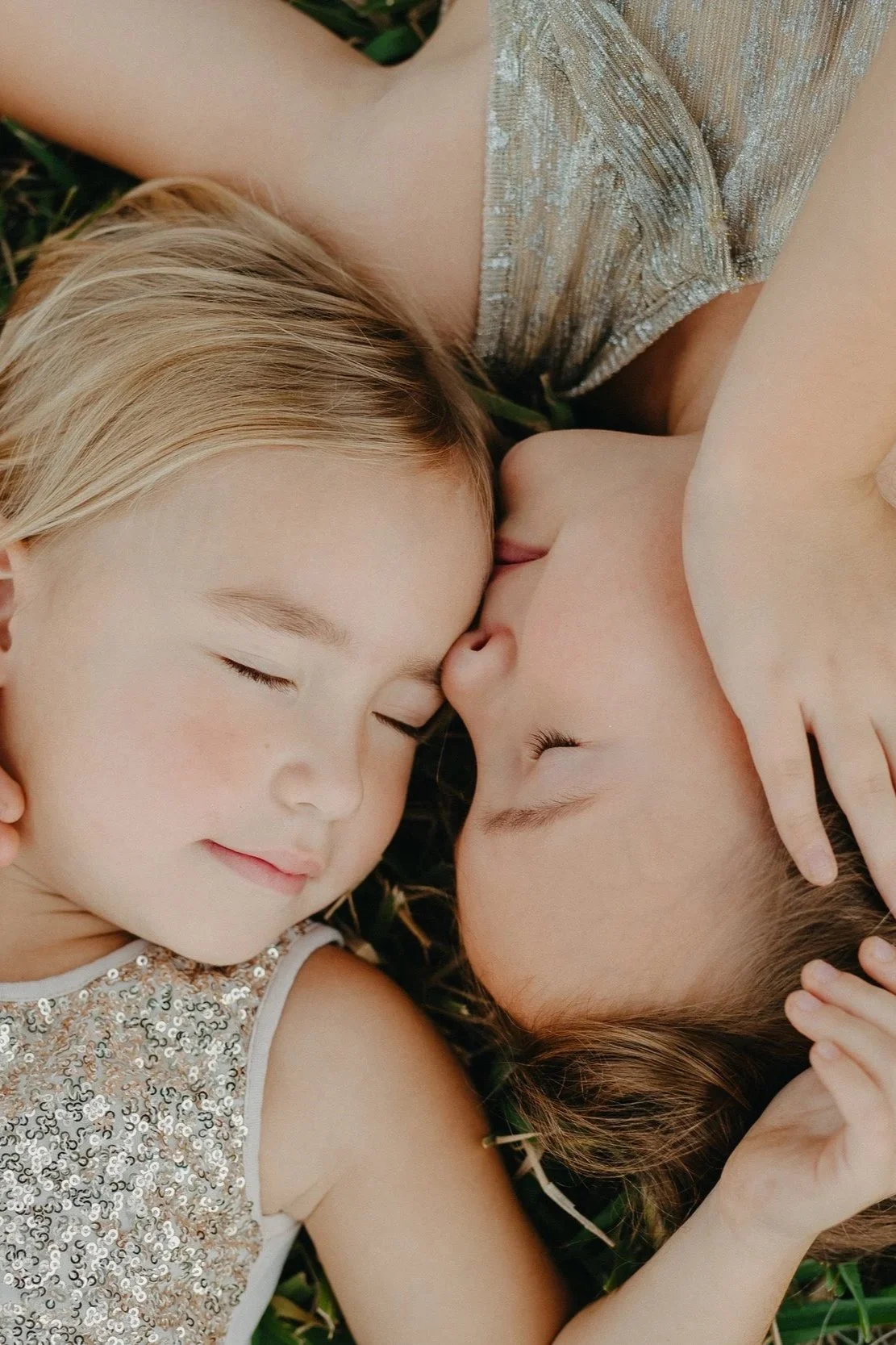 Two young girls with closed eyes lying on grass, touching foreheads gently and smiling peacefully.