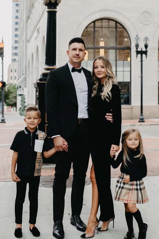 Family of four in formal attire standing outdoors in an urban setting, holding hands, with historic building and street lamps in the background.
