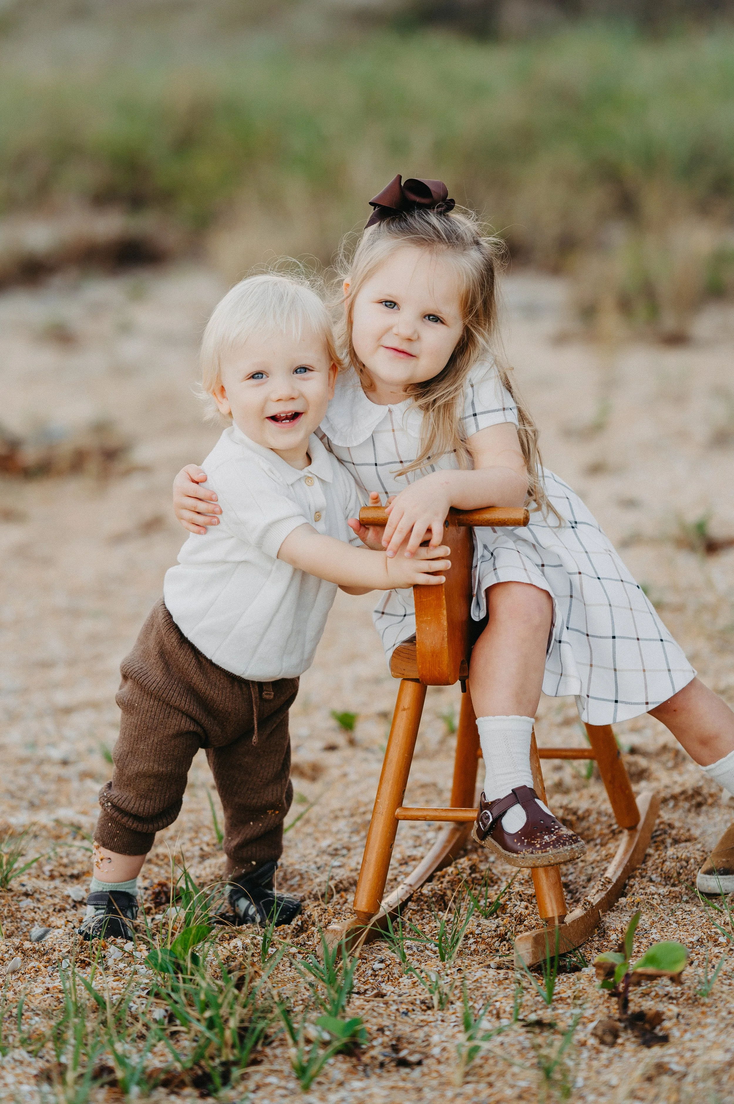 A young girl and a toddler boy outdoors on a sandy area with some green plants, smiling and holding onto a wooden rocking horse.