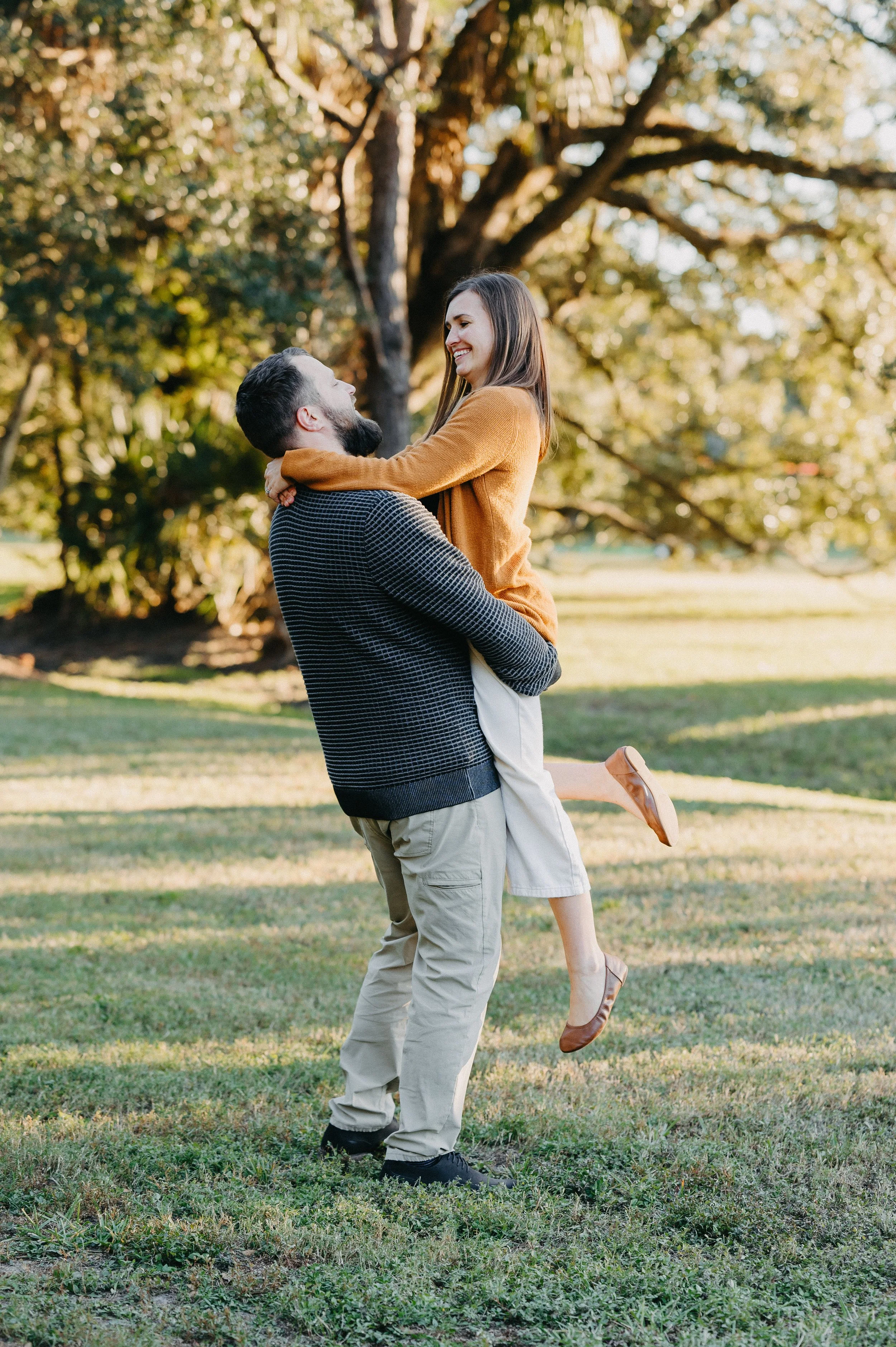 A man lifting a woman in a park with trees in the background, both smiling and looking at each other.