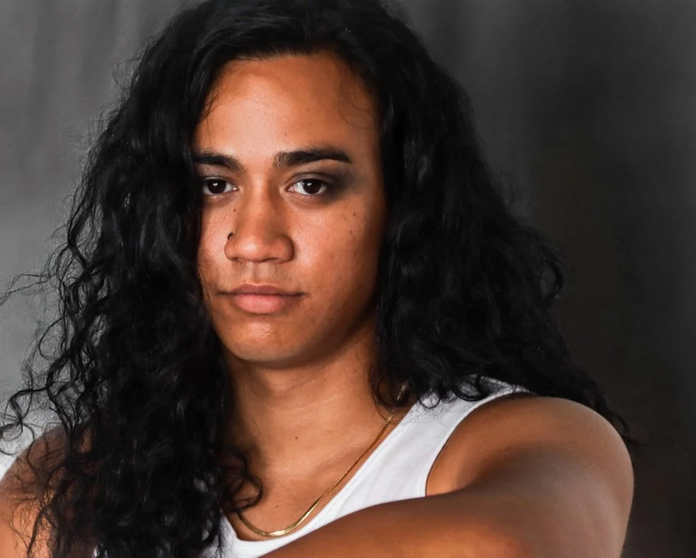A woman with long, curly black hair, medium skin tone, wearing a white sleeveless top and a gold necklace, looking directly at the camera against a dark background.