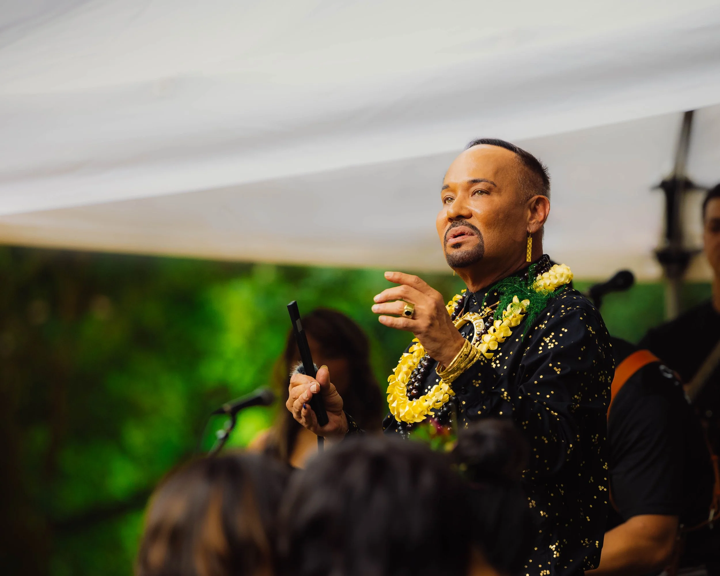 A man speaking at an outdoor event, wearing a black shirt with gold specks, yellow flower lei, and jewelry.