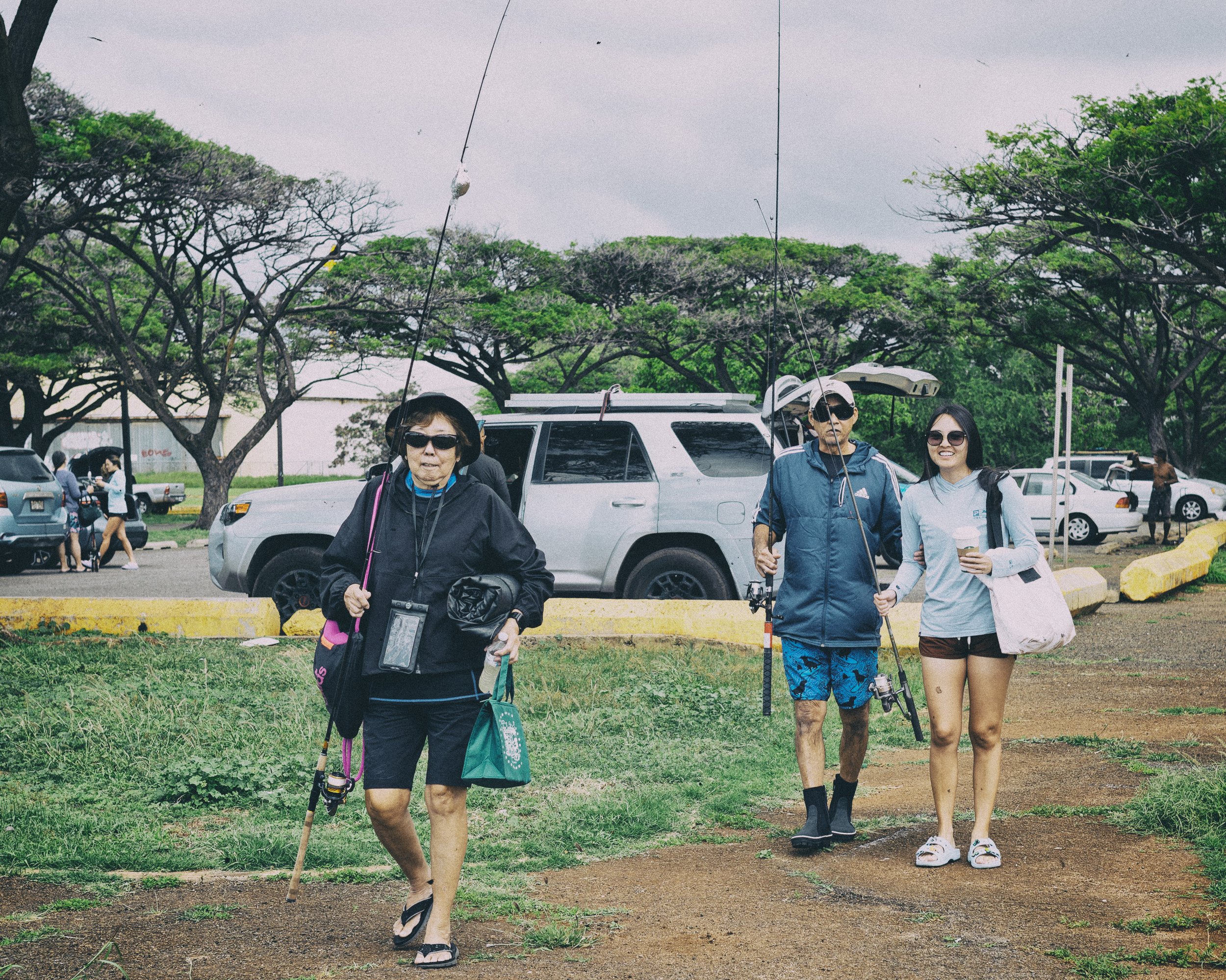 Three people walking outdoors near parked cars, some holding fishing rods, with trees in the background and a cloudy sky.