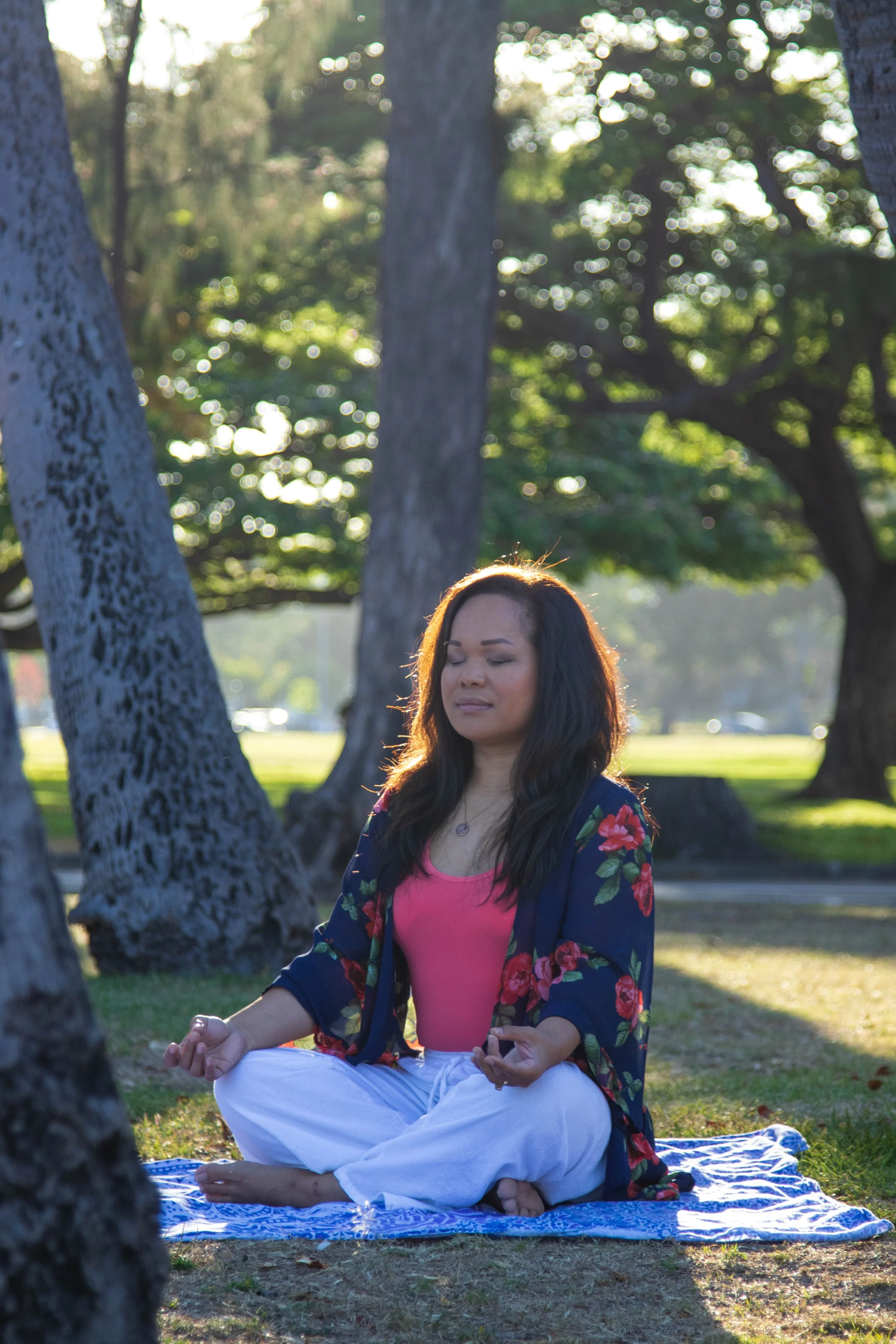 A woman sitting cross-legged on a patterned blue and white blanket in a park, practicing meditation with her eyes closed. She is wearing a pink top, white pants, and a navy blue floral kimono. The background features large trees and sunlight filtering through the leaves.