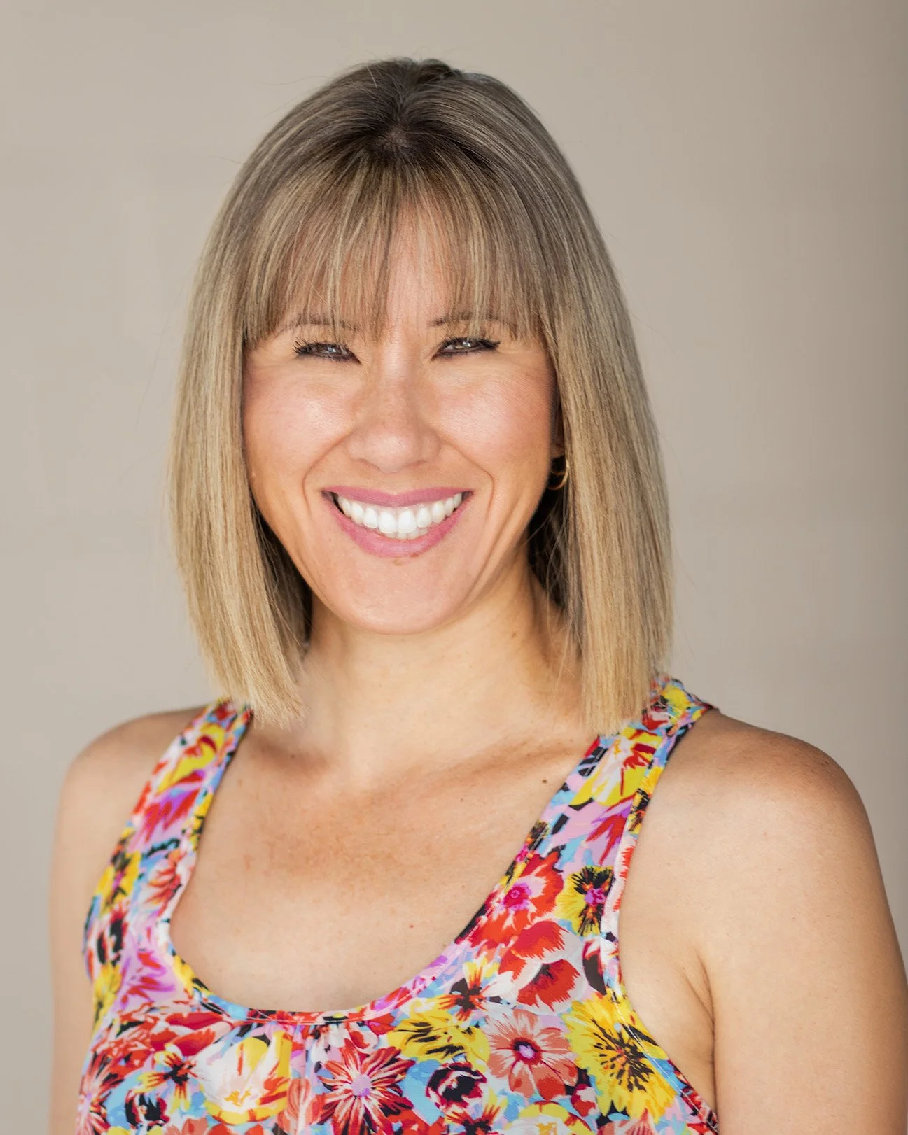 Smiling woman with shoulder-length light brown hair, wearing a colorful floral sleeveless top, against a plain neutral background.