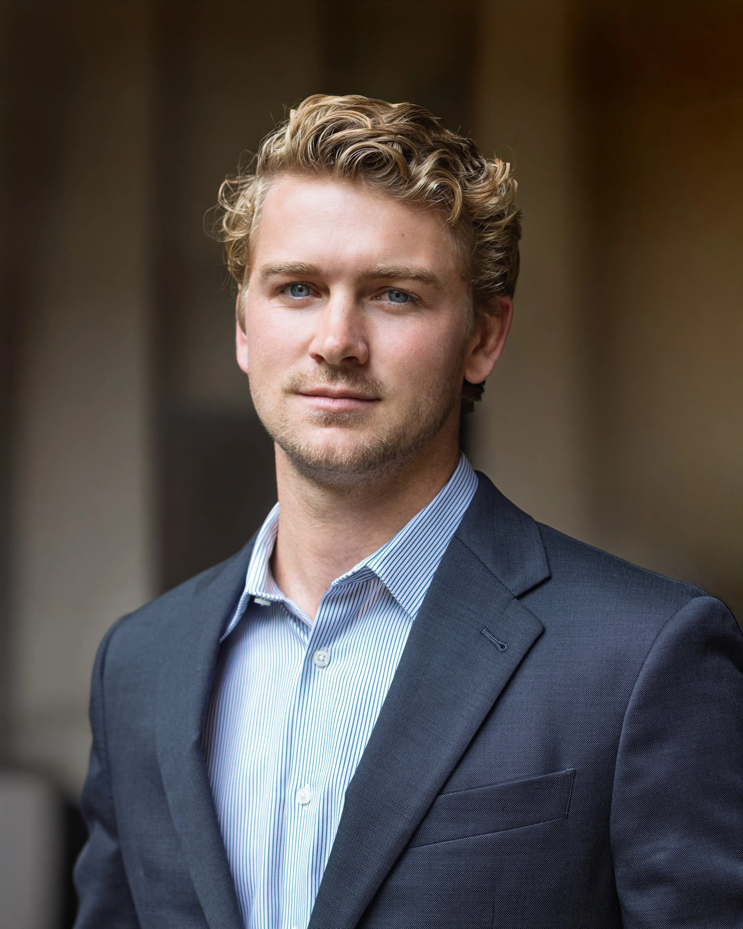 A young man with blue eyes, blond curly hair, wearing a dark suit and a light blue striped shirt, standing outdoors with a blurred background.