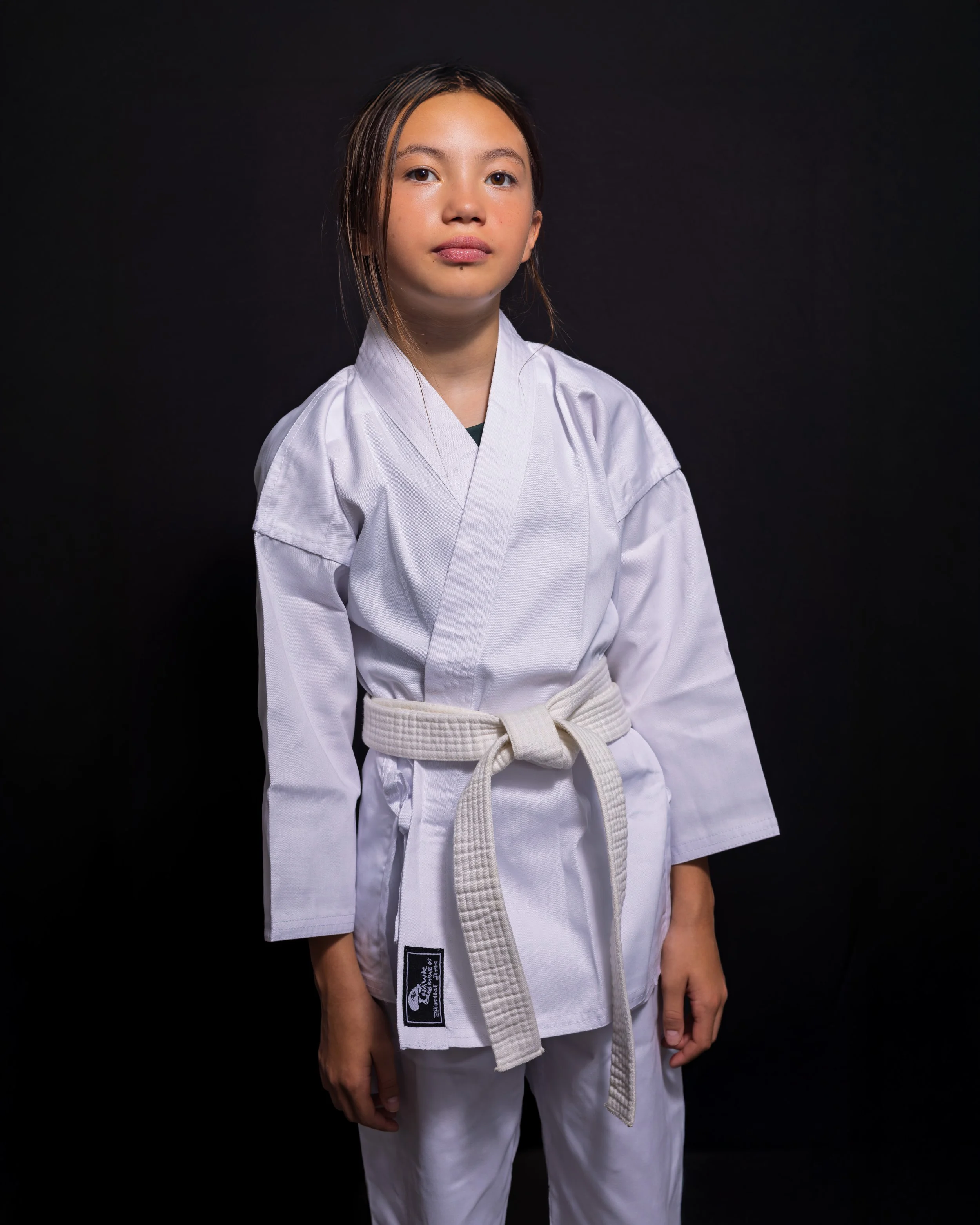 Young girl wearing a white karate gi with a beige belt standing against a black background.