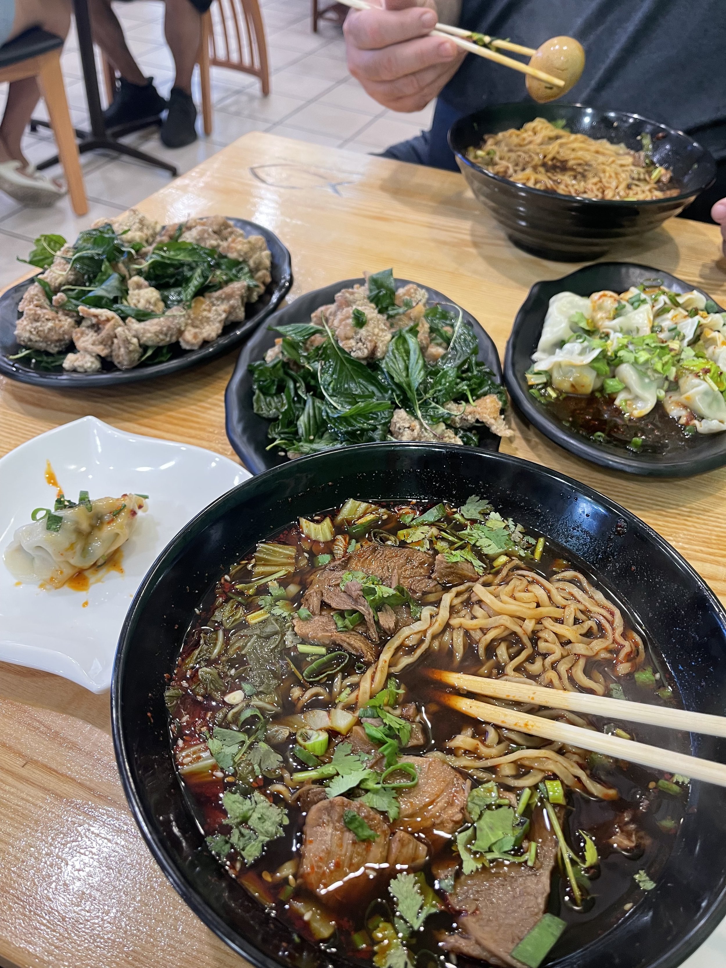A spread of Asian dishes at a restaurant, including bowls of noodles, dumplings, fried greens, and a small plate of dumplings with chili sauce on a wooden table, with a person handling chopsticks.