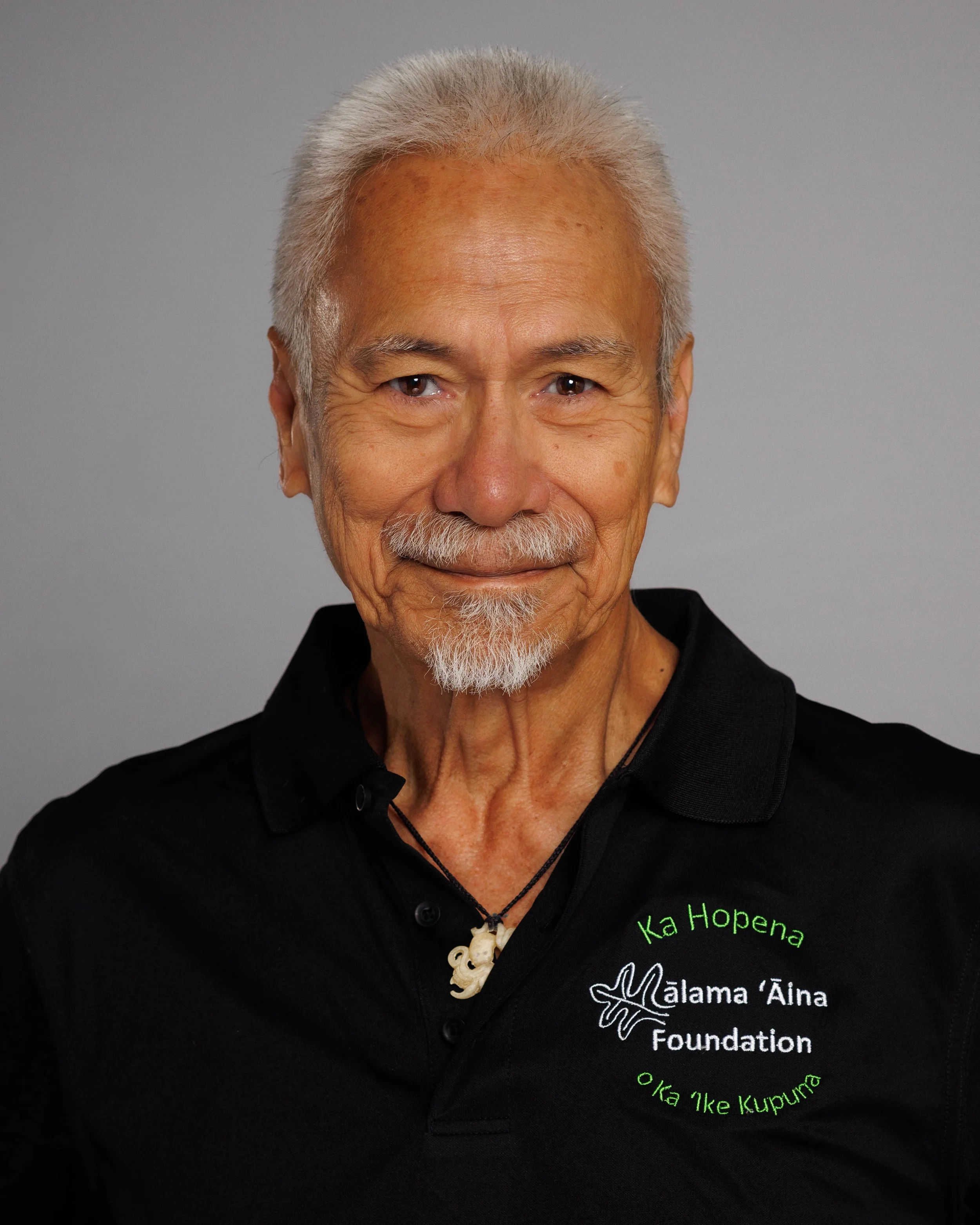 Portrait of an elderly man with gray hair and a goatee, smiling, wearing a black polo shirt with green and white embroidered logo and text, and a necklace with a pendant, against a plain gray background.