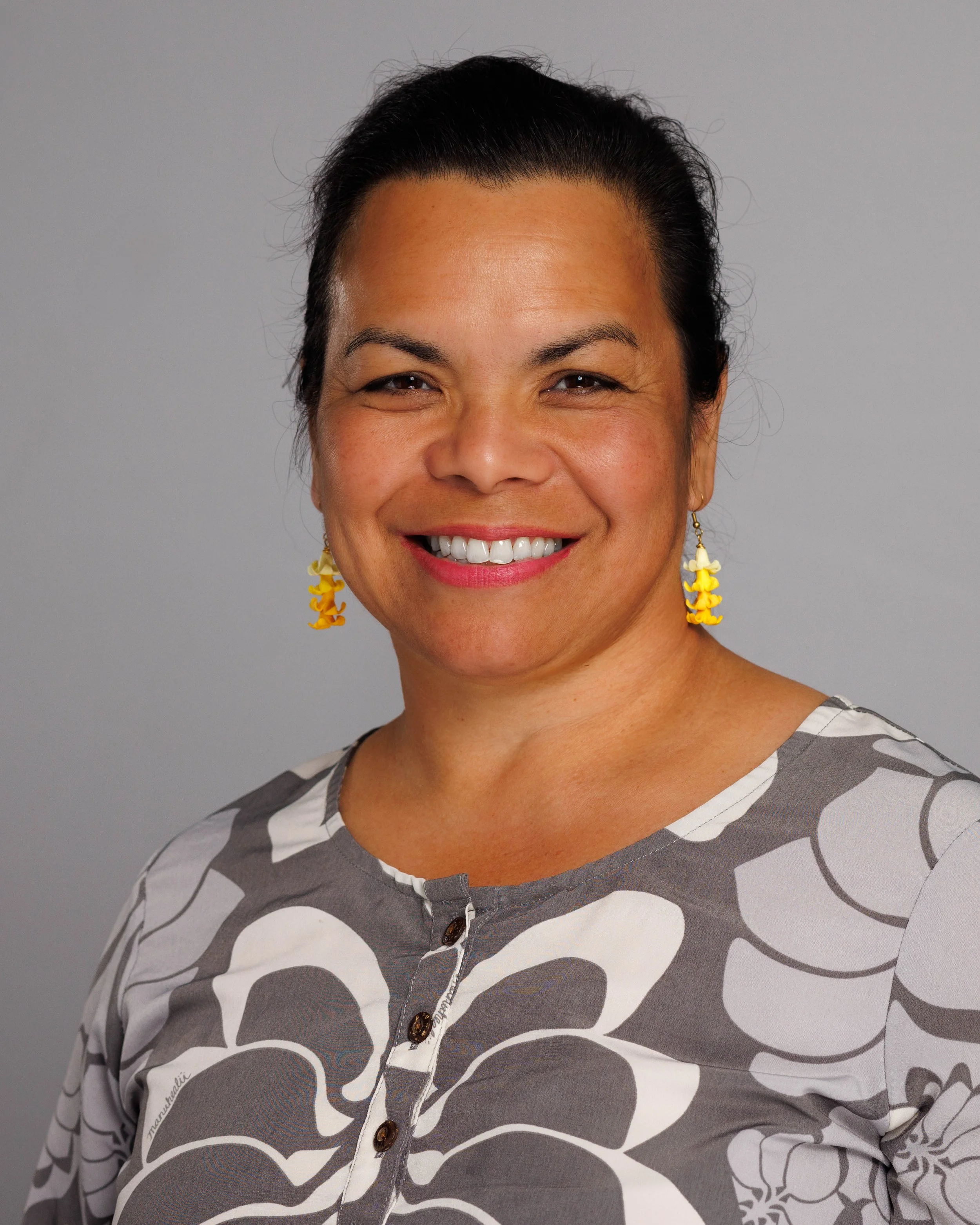 Woman with dark hair pulled back, smiling, wearing yellow floral earrings and a gray and white patterned blouse, standing against a plain gray background.