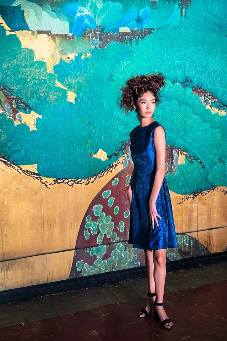 A young woman with curly hair stands against a colorful mural of a dragon painted on a wooden wall. She is wearing a sleeveless, knee-length, blue patterned dress and black high-heeled sandals.