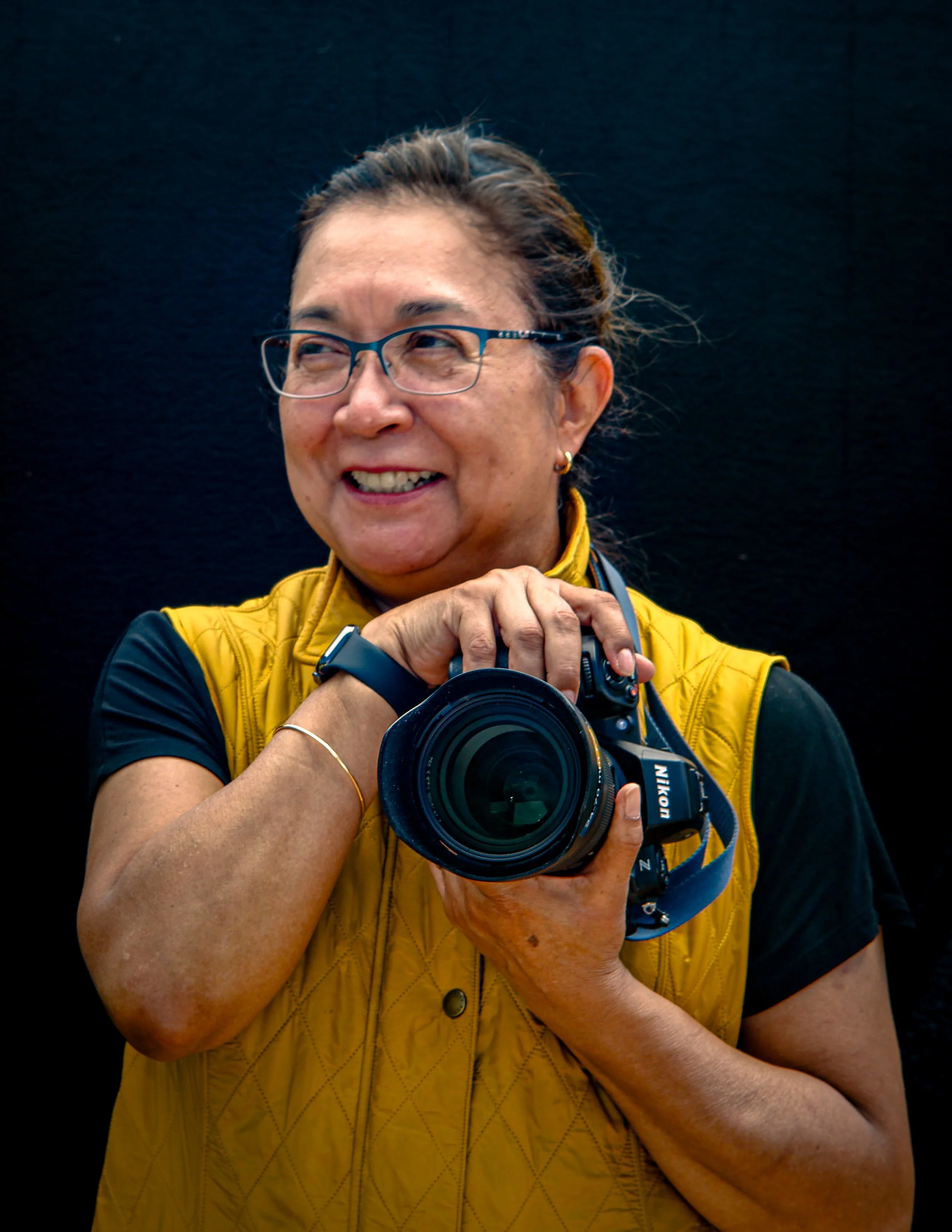 Woman with glasses, wearing a yellow vest and black shirt, holding a Nikon camera, smiling in front of a black background.