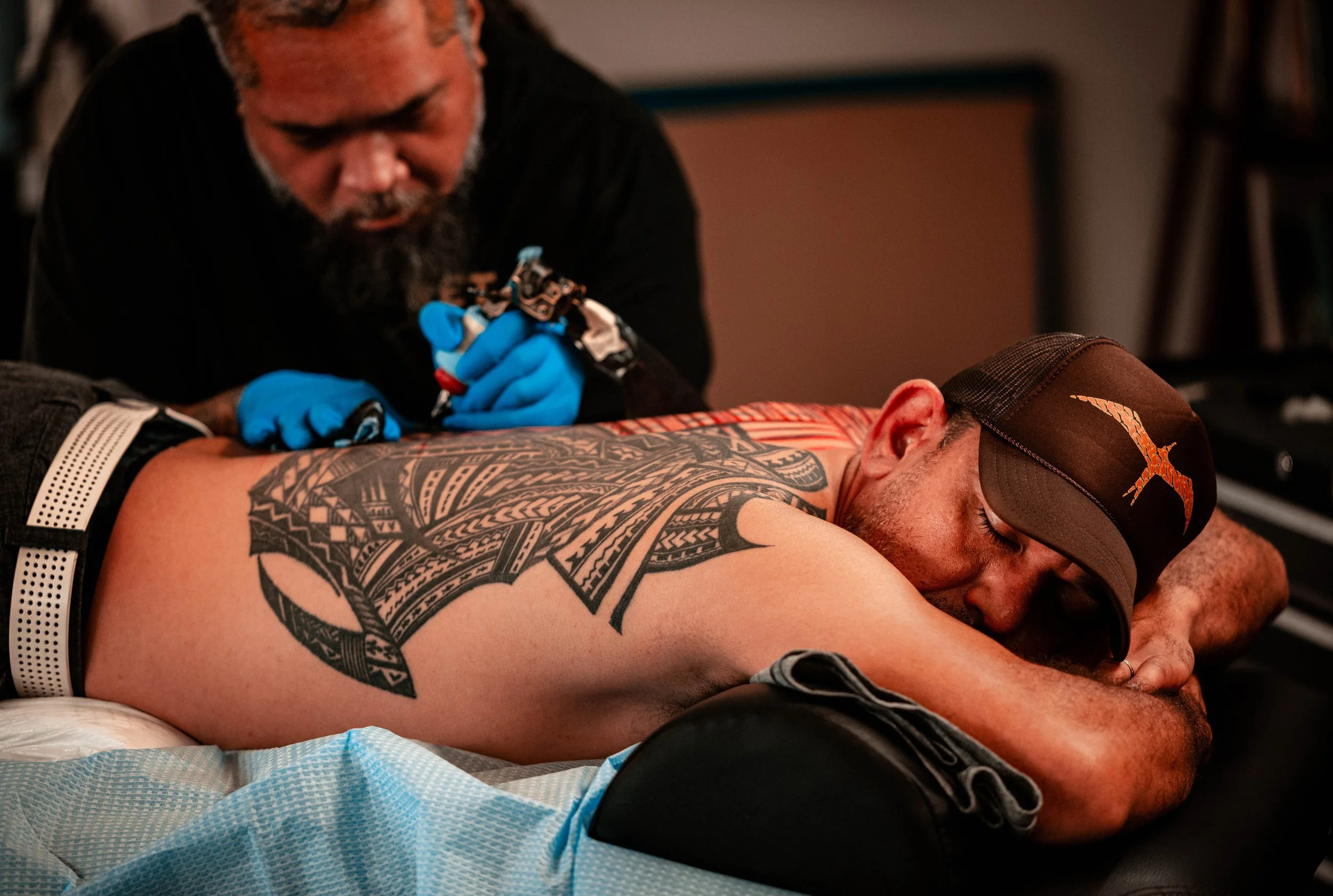 A man with a large tribal tattoo on his back receives a tattoo from an artist wearing blue gloves.