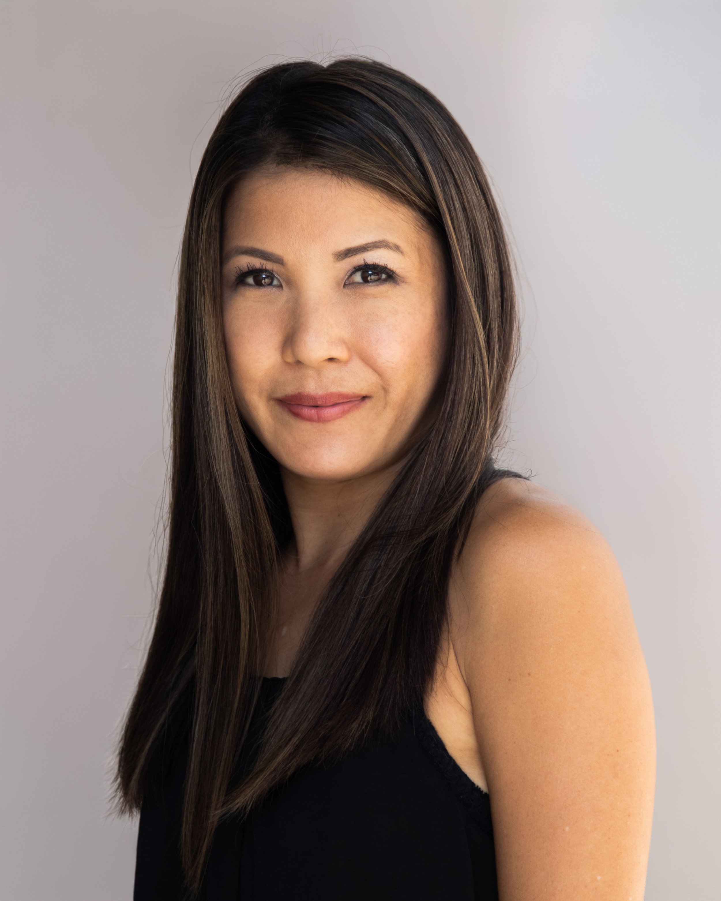 Close-up portrait of a woman with straight, dark hair, wearing a black sleeveless top, smiling softly against a plain light background.