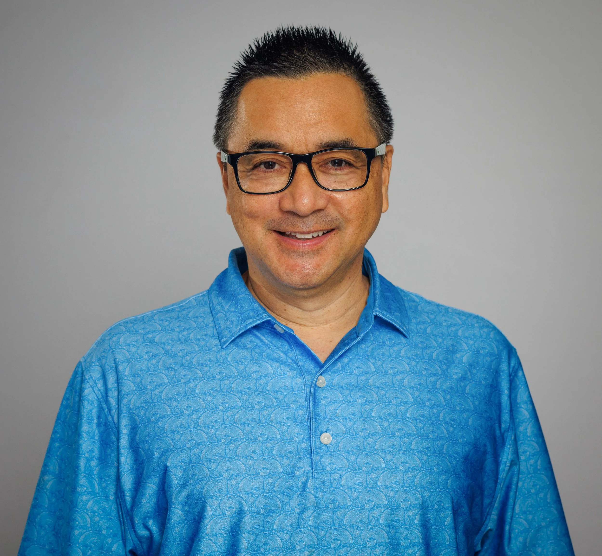 A smiling man with short black hair, wearing glasses and a blue patterned button-up shirt, standing against a plain gray background.