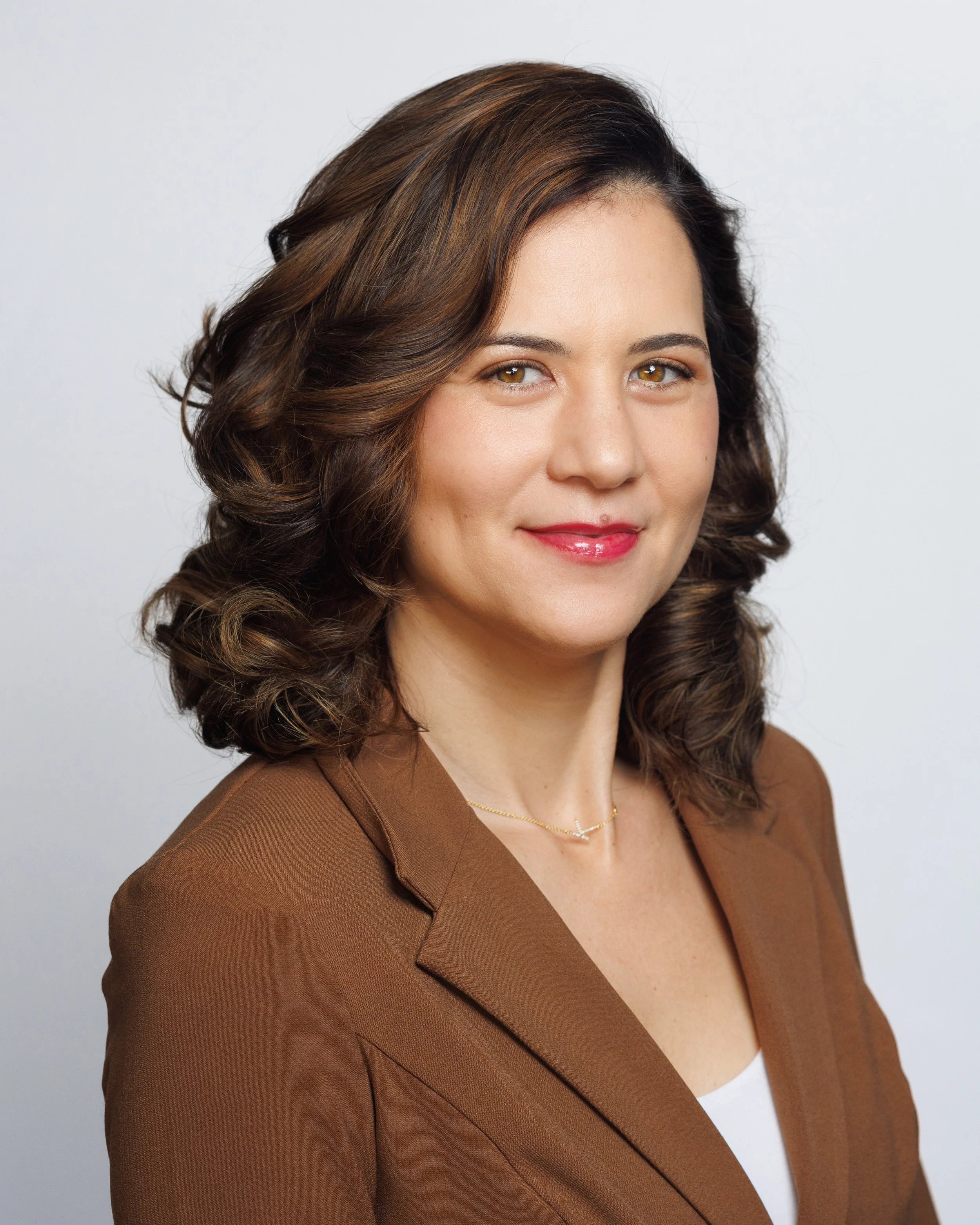 A professional woman with brown, curled hair wearing a brown blazer and smiling