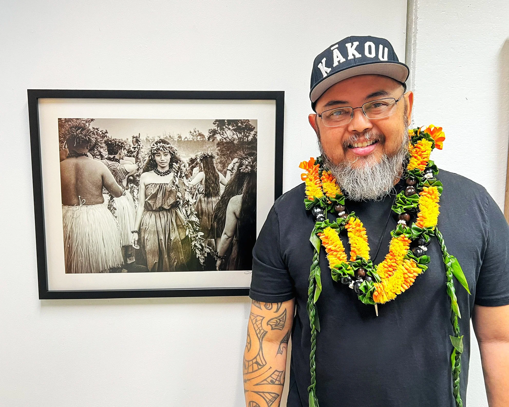 A smiling man with glasses and a gray beard wearing a black T-shirt, a khaki cap with the words 'KĀKOU', and multiple colorful leis, standing in front of a framed black-and-white photo of people in traditional attire dancing.