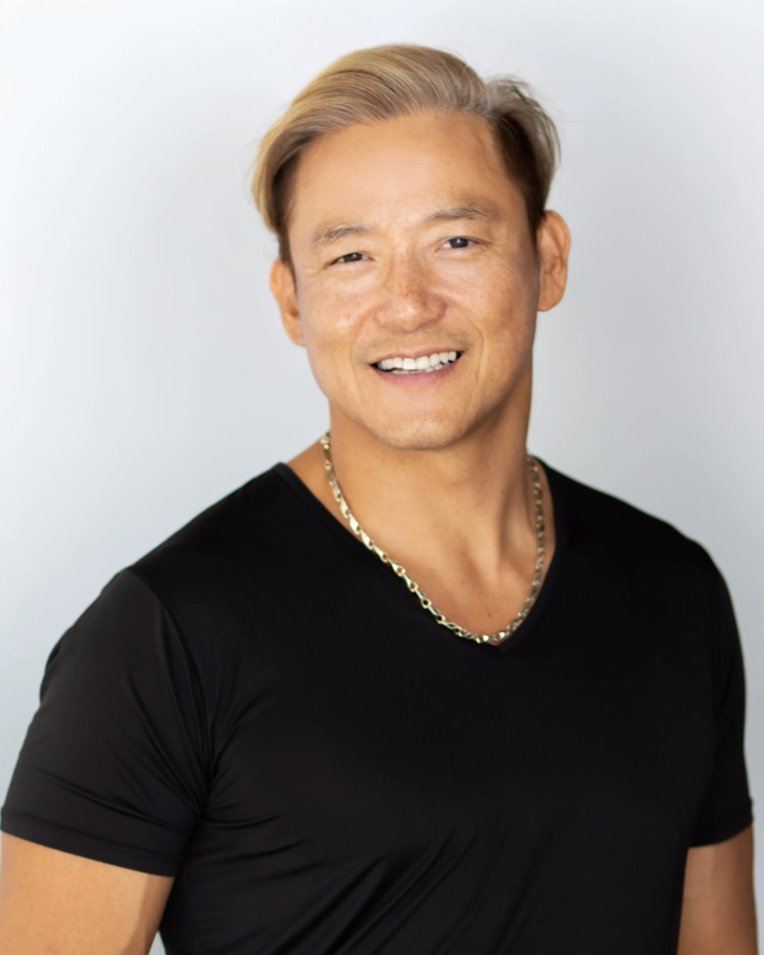 Portrait of a smiling man with blonde hair in a black shirt and gold chain necklace, against a plain white background.