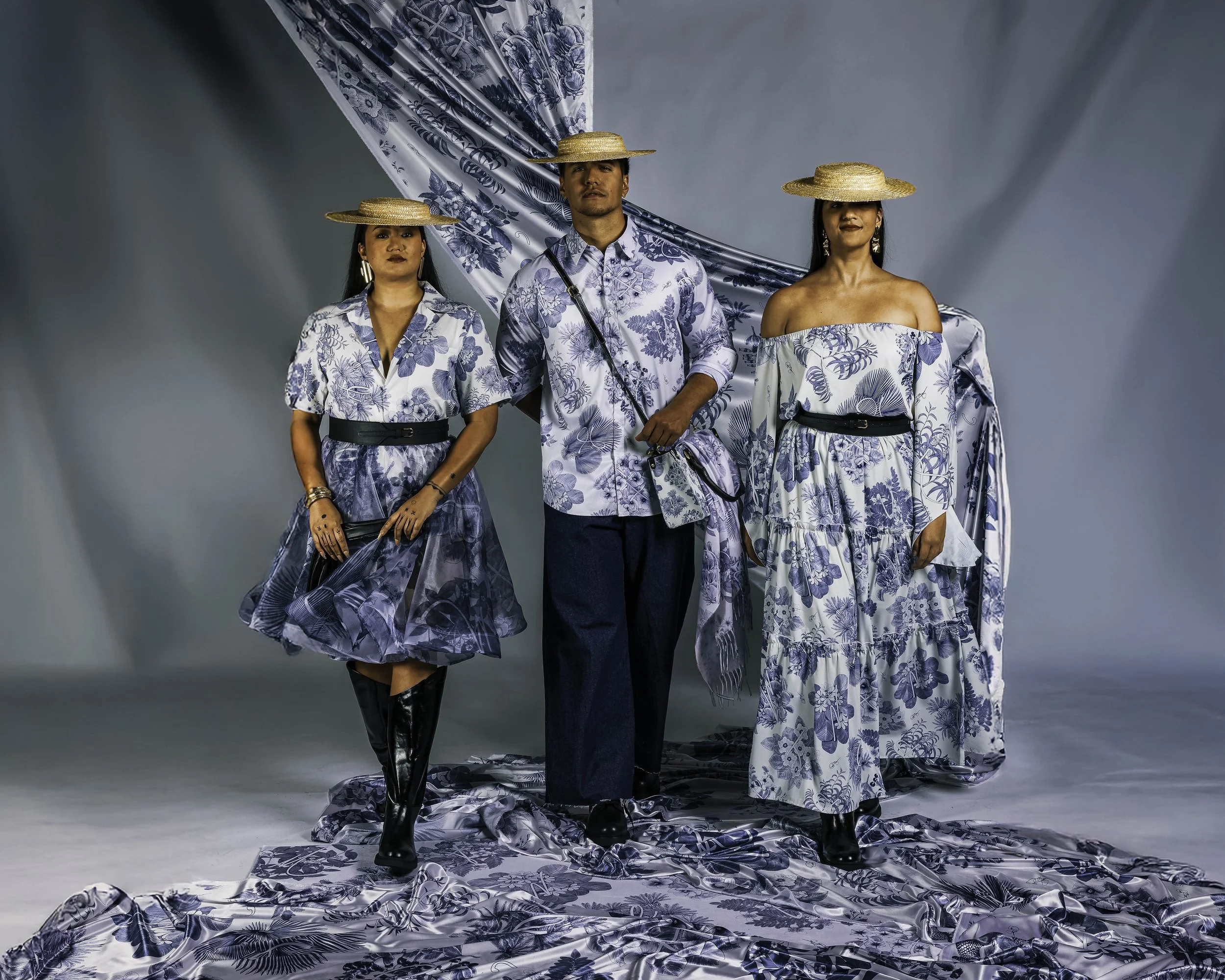 Three people dressed in blue and white floral outfits wearing straw hats standing on a floral fabric backdrop.