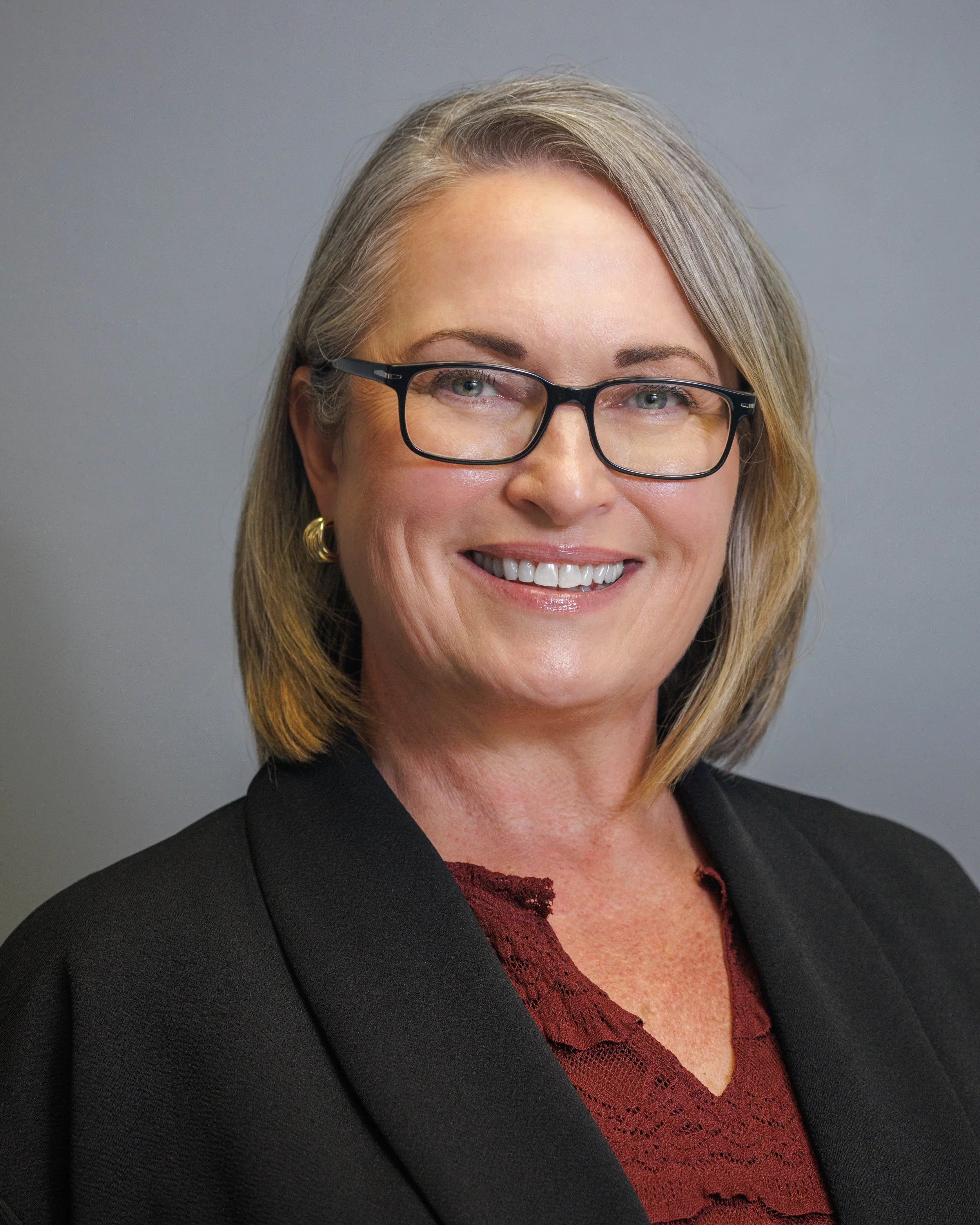 Professional woman with shoulder-length gray hair, glasses, wearing a black blazer and burgundy lace top, smiling against a gray background.