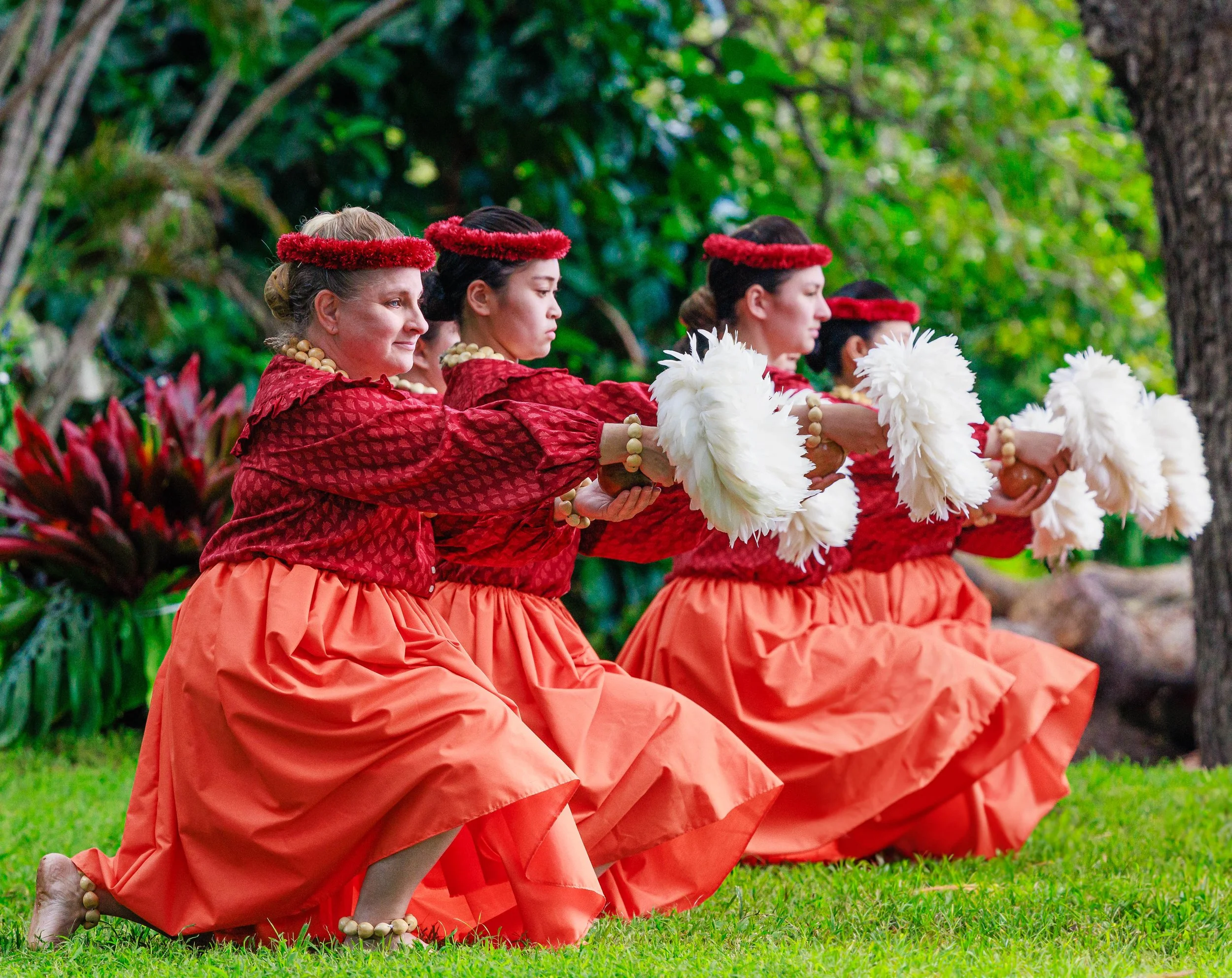Four women dressed in traditional Polynesian attire, kneeling on grass, holding white feathered fans, with greenery in the background.
