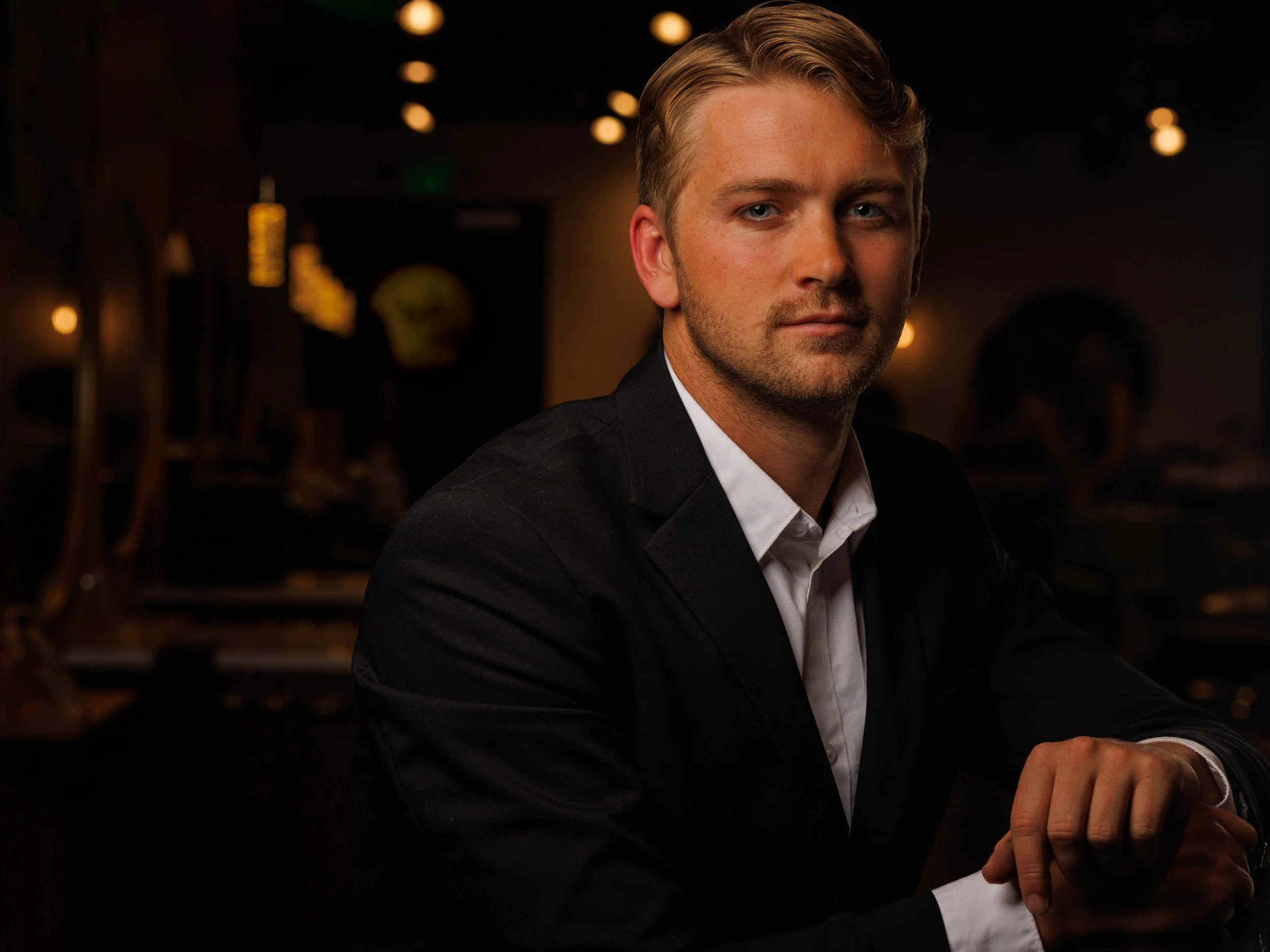 A man with blonde hair and a light beard, wearing a black suit and white shirt, seated in a dimly lit restaurant or bar with warm lighting.