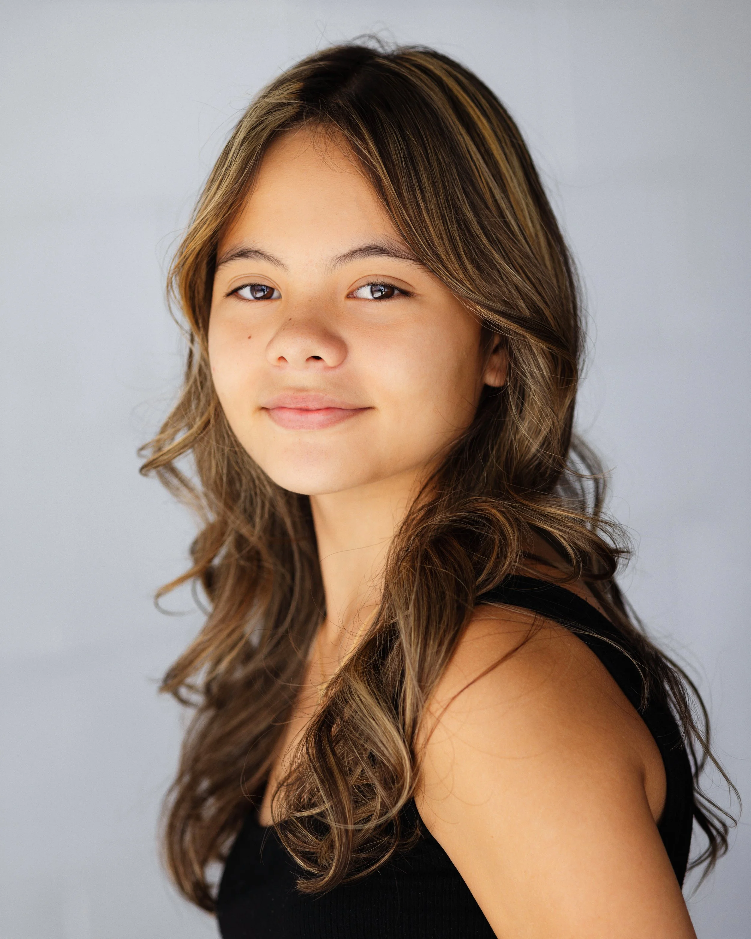 Close-up portrait of a young woman with long wavy brown hair, wearing a black tank top, smiling softly against a plain light gray background.