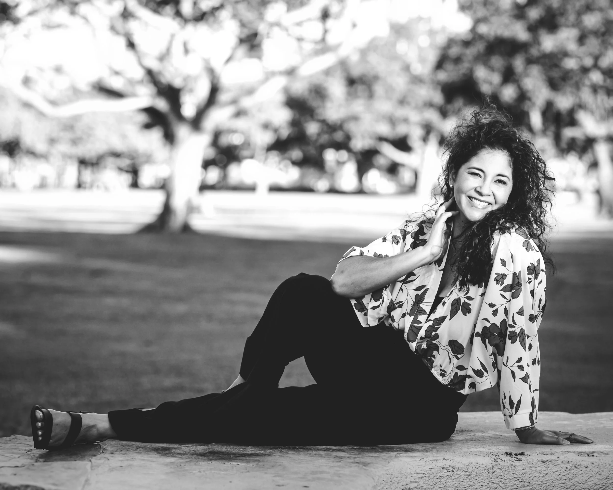 A woman with curly hair, smiling and winking, sitting on a low stone wall in a park with trees in the background.