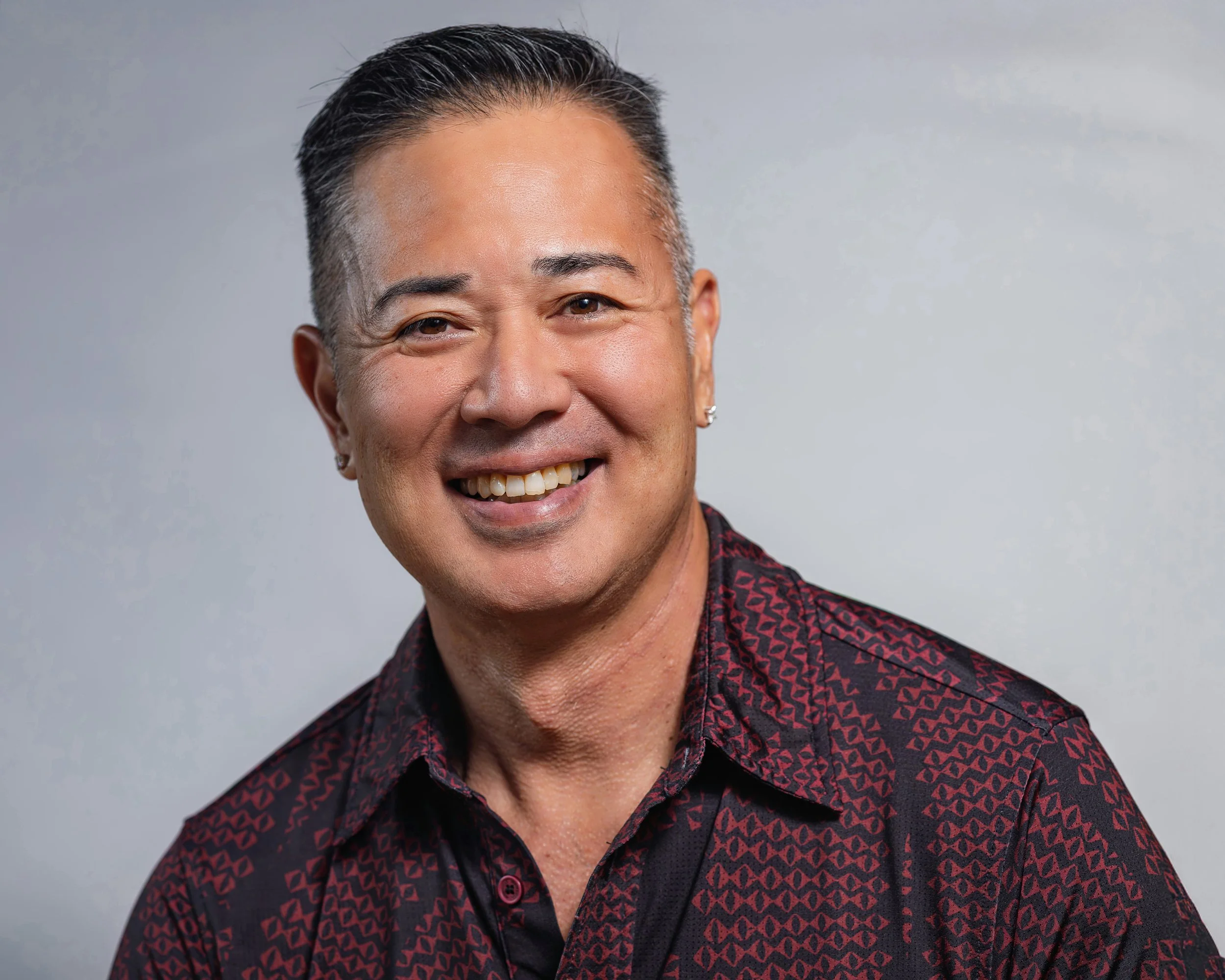 Headshot of a smiling middle-aged man with short dark hair, wearing earrings, and a patterned dark red and black shirt against a plain background.