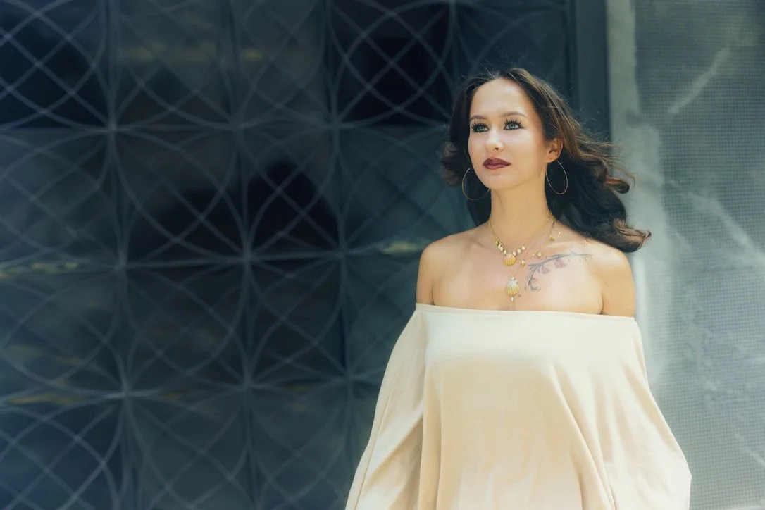 A woman with dark wavy hair and hoop earrings stands outdoors, wearing an off-the-shoulder cream dress and layered gold jewelry, with a dark modern building in the background.