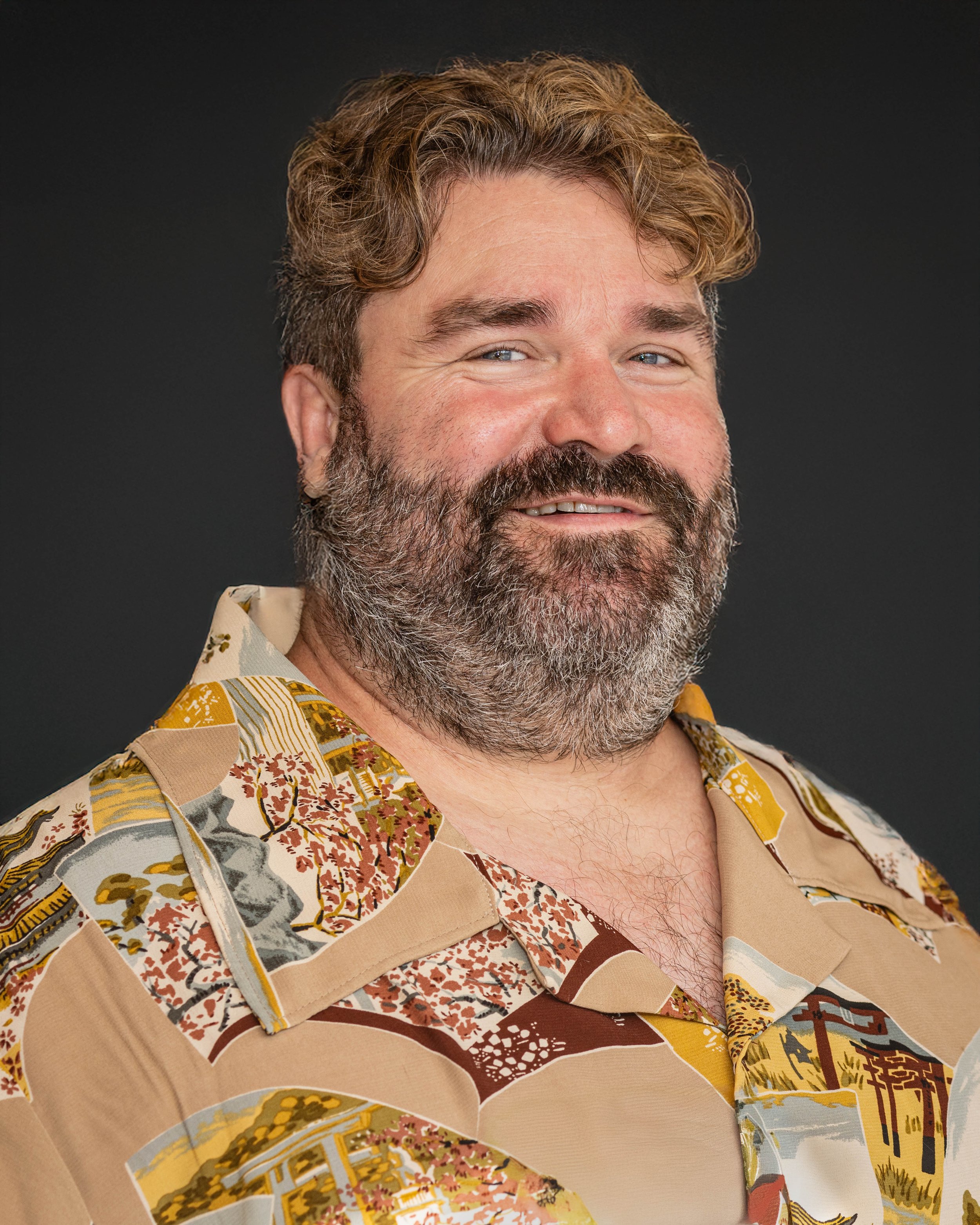 A middle-aged man with a beard and wavy hair smiling, wearing a colorful hawaiian shirt, against a dark background.