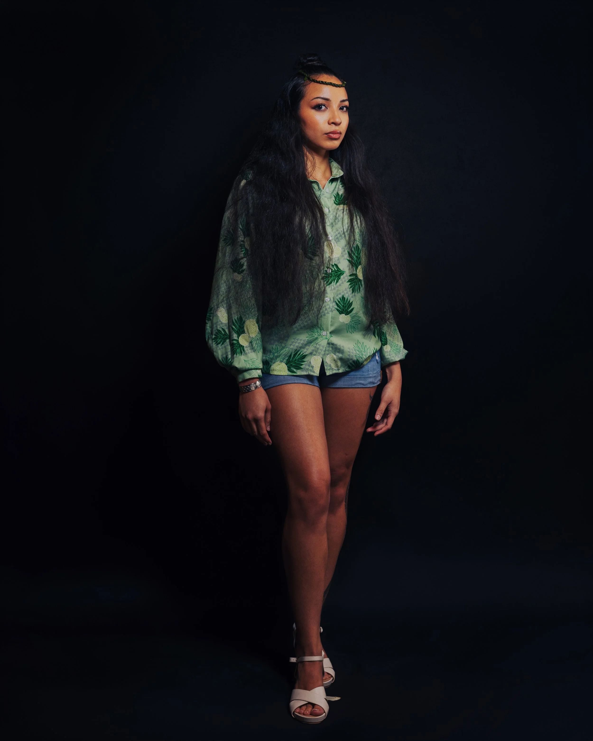 A woman with long, dark, wavy hair, wearing a green tropical print blouse, jean shorts, and white high heels, standing against a black background.