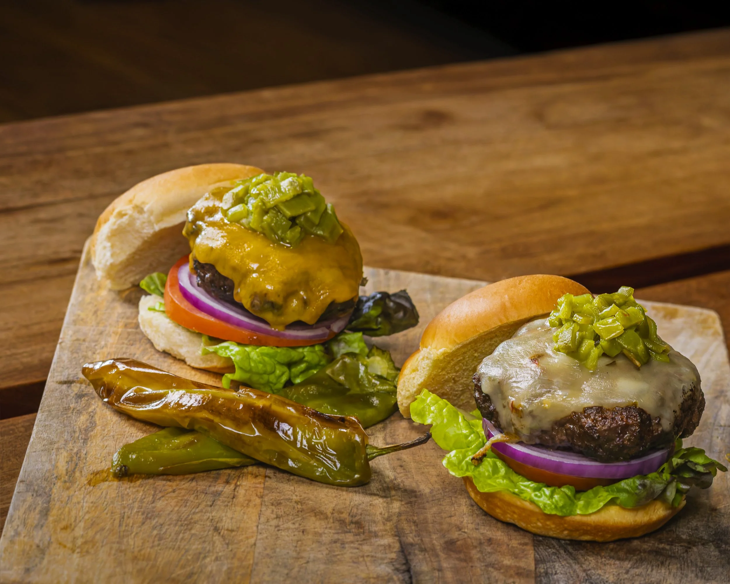 Two cheeseburgers with lettuce, tomato, onion, pickles, and melted cheese, topped with chopped pickles, served on a wooden board with roasted chili peppers.