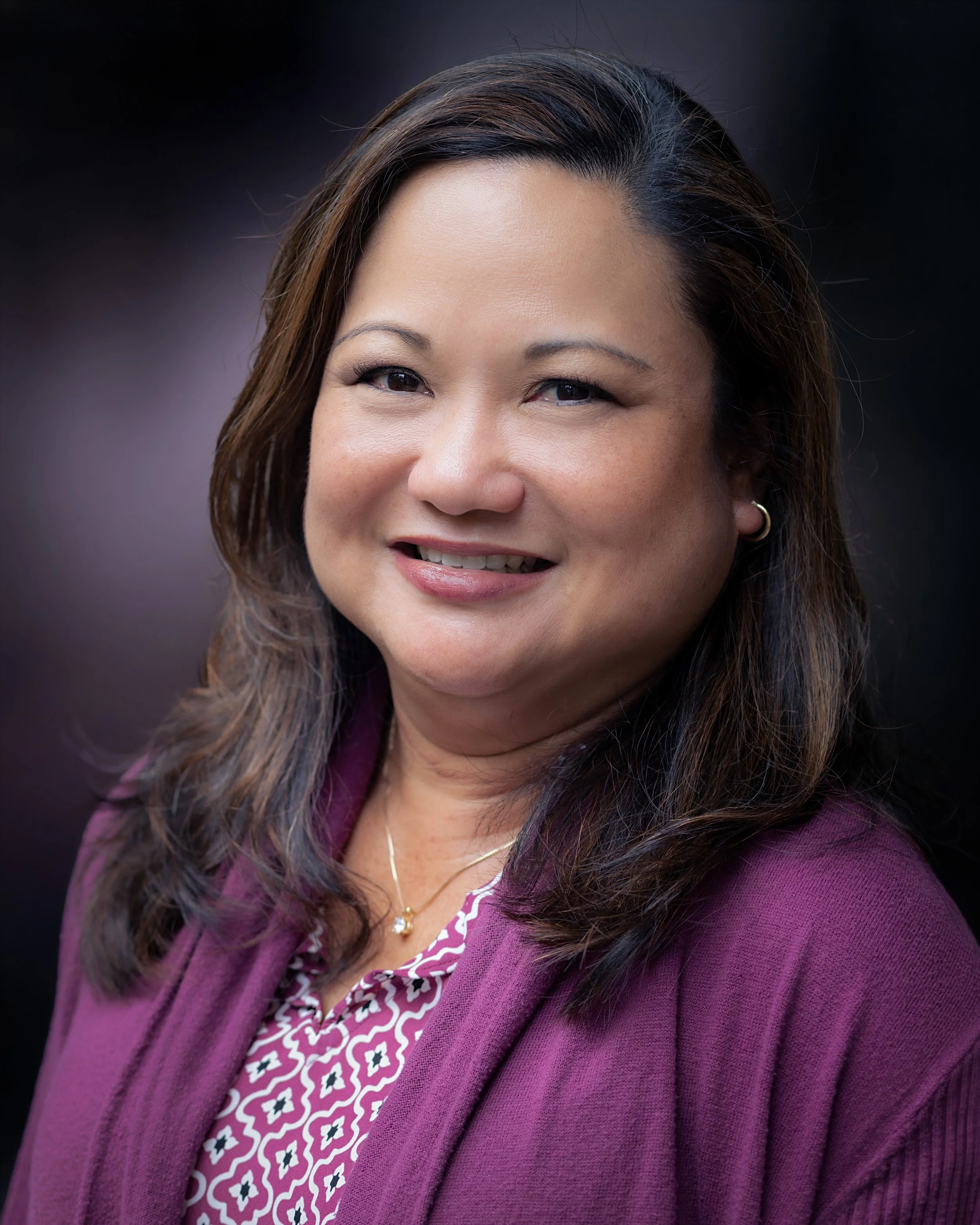 A professional woman with medium-length dark hair, wearing a patterned blouse and a purple jacket, smiling against a dark background.
