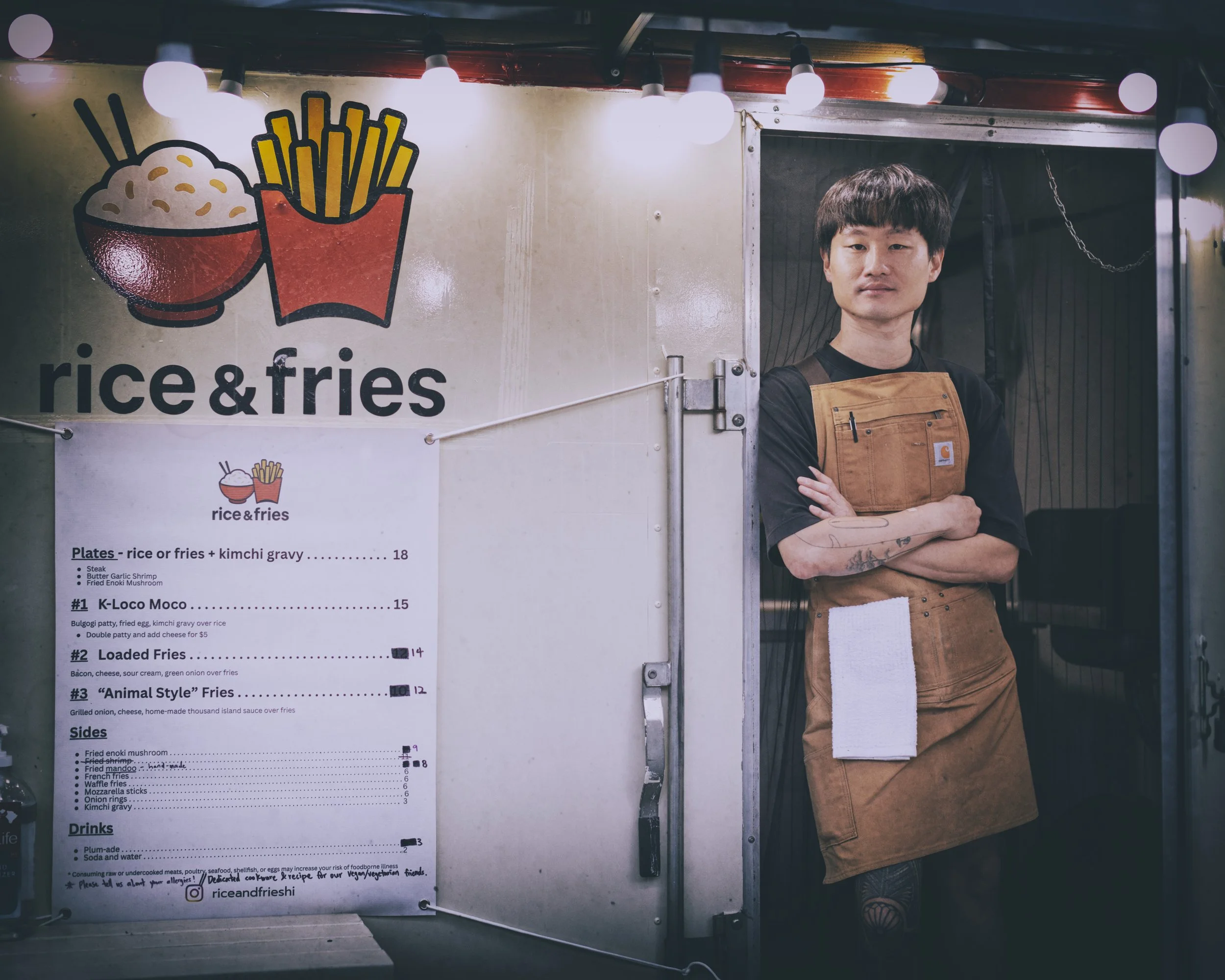 A man standing in a food stall with a sign that reads 'rice & fries', and a menu listing various rice and fries dishes, wearing a brown apron and black shirt.