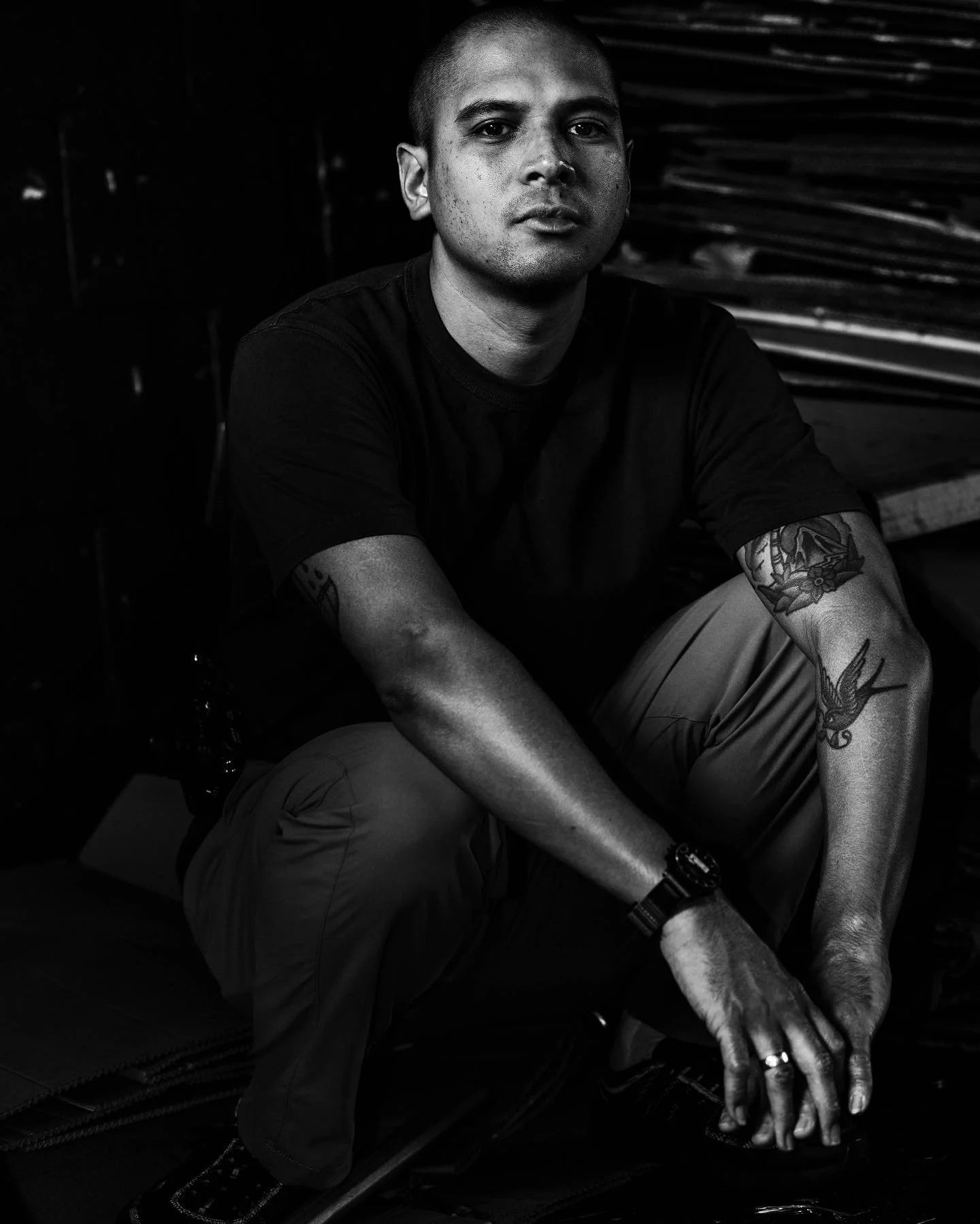 A black and white photo of a young man with short hair, tattoos on his arms, wearing a black T-shirt, sitting with his hands clasped, looking directly at the camera, with a dark background.