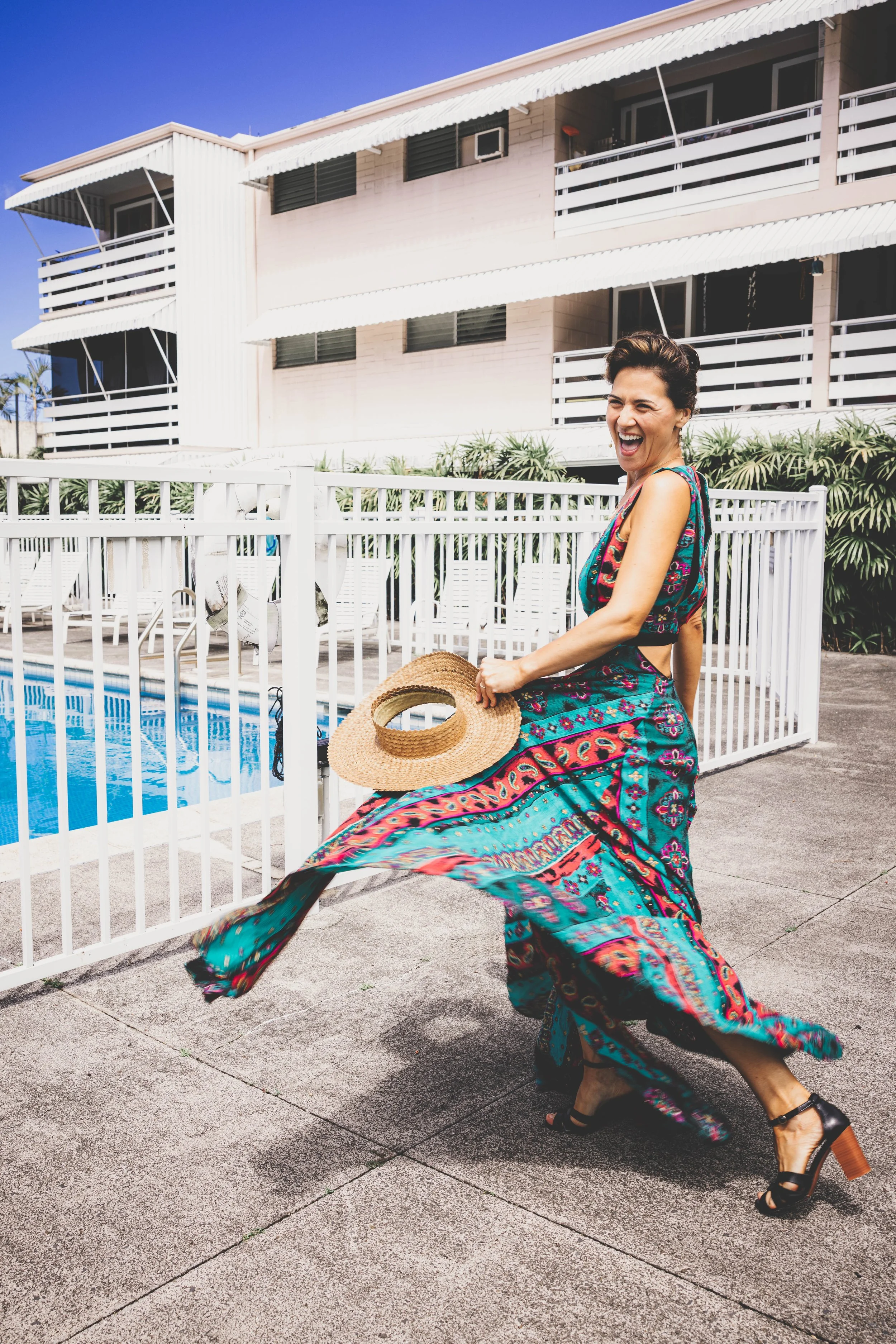 Woman in colorful dress holding a straw hat, smiling and twirling outside near a swimming pool with apartment buildings in the background.