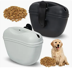 Two small black and white portable dog food containers, one with a pet dog sitting nearby, and two piles of kibble, one in the foreground and one in the top left corner.