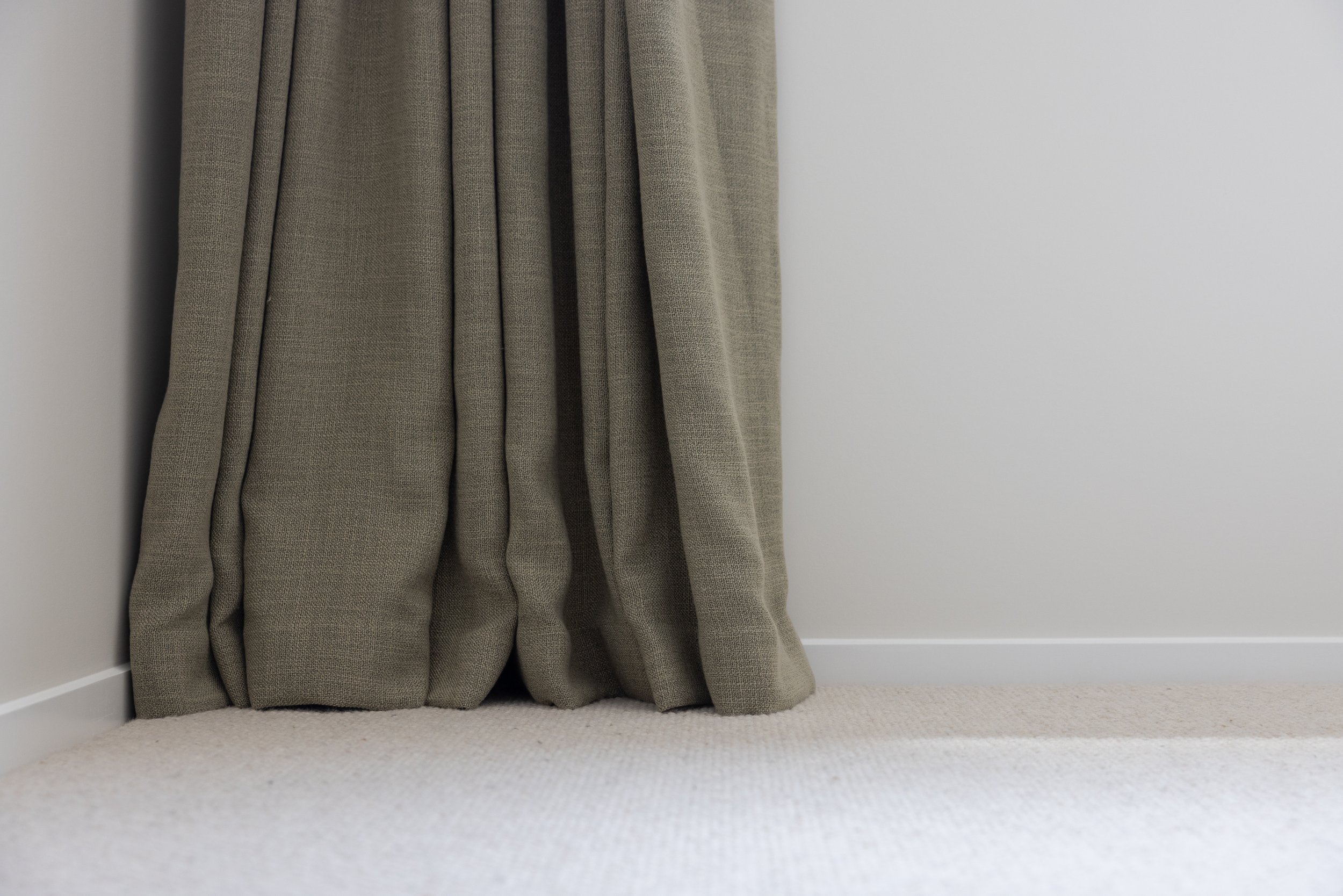 Close-up of beige curtains hanging next to a white wall and beige carpeted floor.