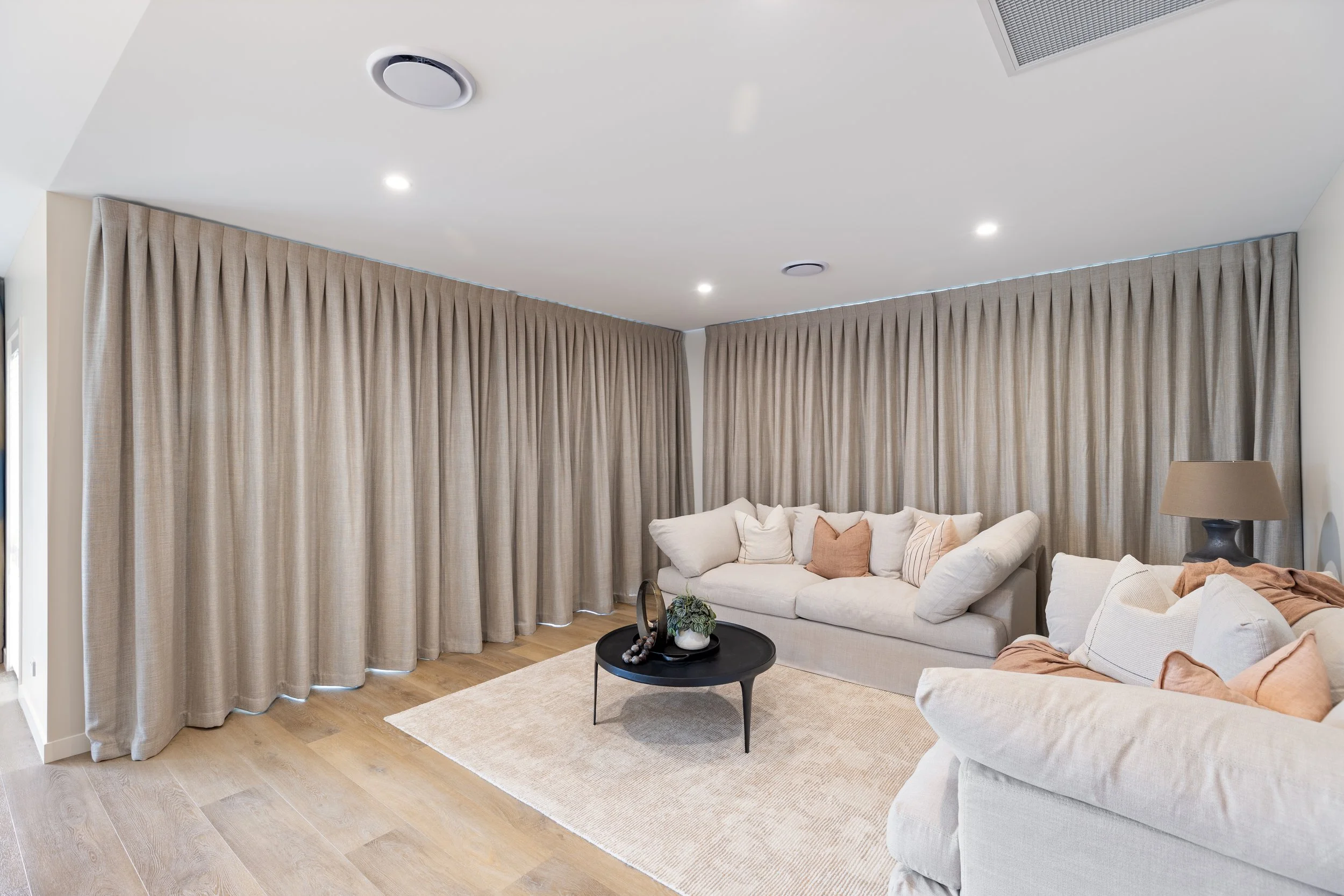 Modern living room with dark gray sofa, white wall-mounted shelves, large windows with gray curtains, and a view of greenery and buildings outside. There is a coffee table with a white throw blanket, cup, and snacks, and unique white side tables near the window.