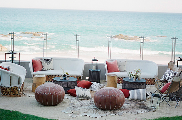 Long outdoor dining table set up with chairs, glasses, and candles along a coastal shoreline at sunset