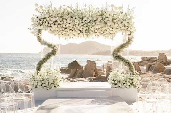 Outdoor dining area with tables and chairs near the beach, surrounded by desert plants, with ocean and distant mountains in the background.
