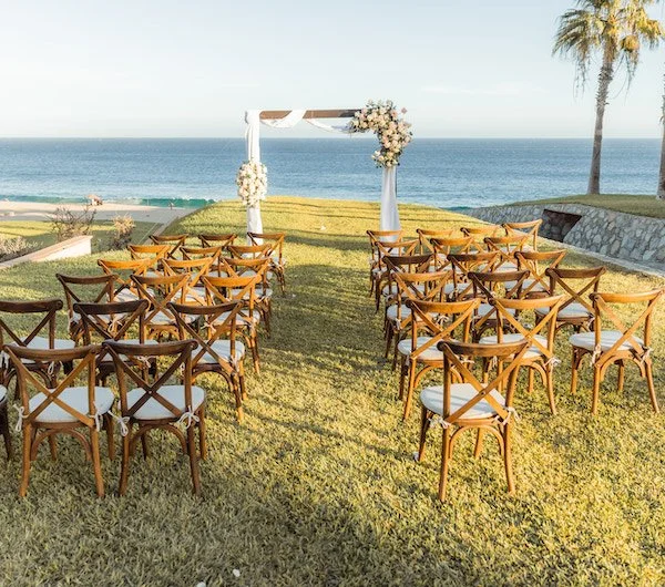 Outdoor wedding setup with wooden chairs facing a floral arch near the ocean, with palm trees and a clear sky.