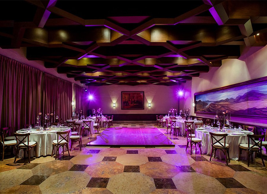 An elegant event hall with round tables covered in white tablecloths, decorated with flowers and glassware, and a dance floor in the center. The ceiling features geometric wooden patterns with purple and yellow accent lighting. The back wall has a large painting and wall sconces, with purple uplighting.