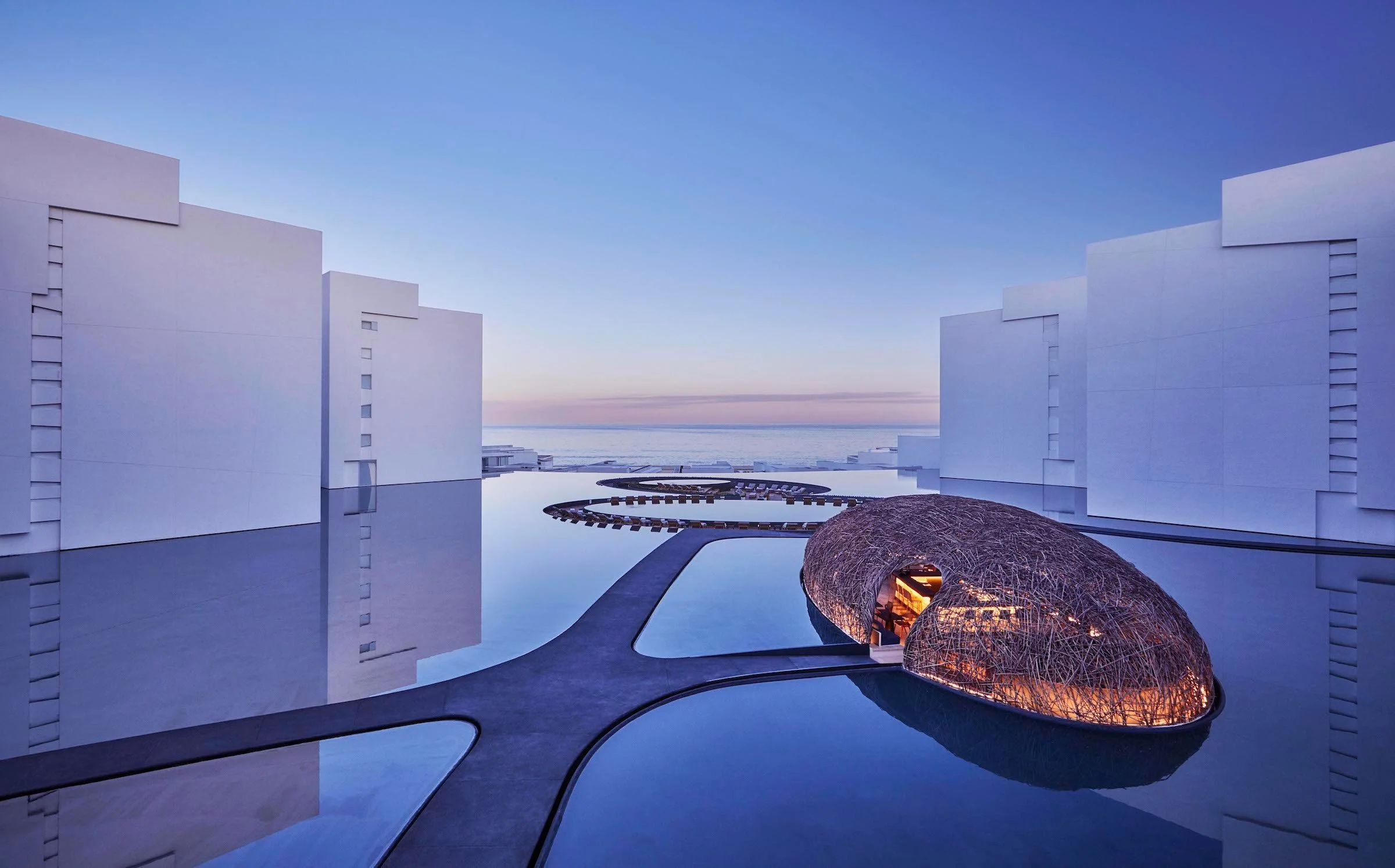 Modern architectural complex with white buildings surrounding a reflective pool, featuring a large, nested egg-shaped structure with an illuminated open section, during twilight with a view of the ocean in the background.
