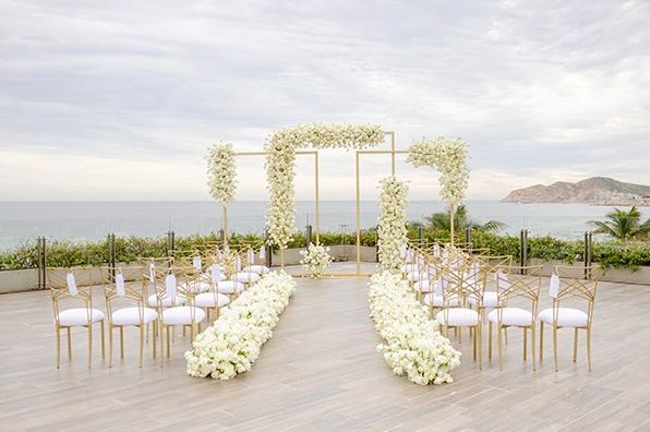 Outdoor patio of a restaurant with wooden tables, white lattice chairs, and large glass windows reflecting sunlight.