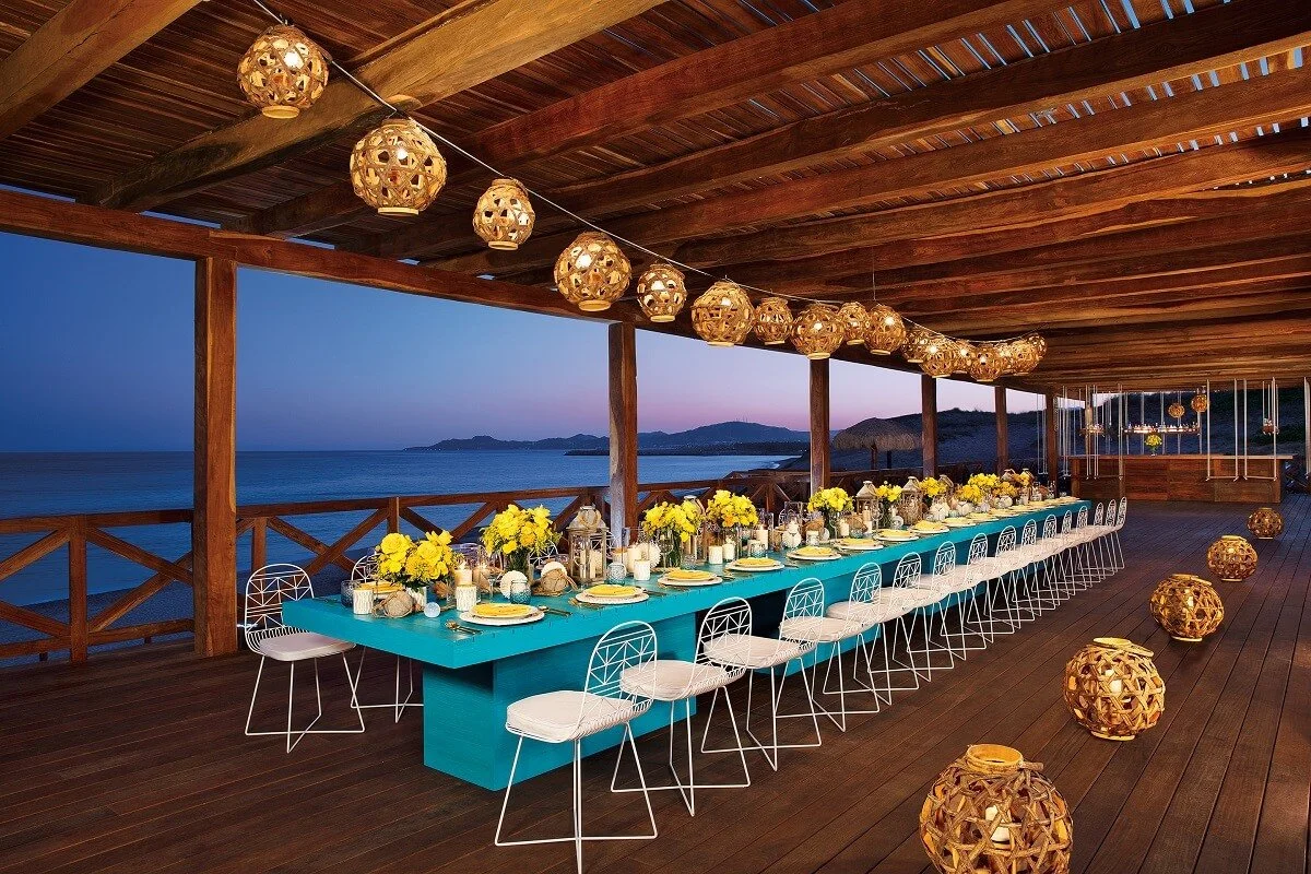 Long outdoor dining table set up with chairs, glasses, and candles along a coastal shoreline at sunset